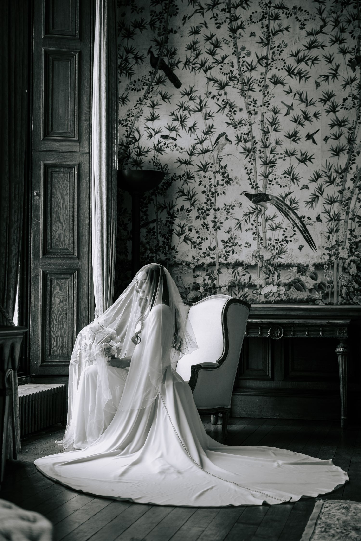 A dramatic black and white bridal portrait featuring a flowing veil and floral wallpaper.