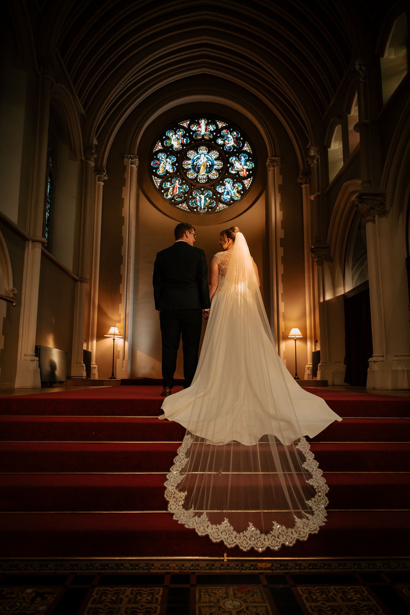 Cathedral wedding photo with ornate stained glass rose window and dramatic veil on red carpet.