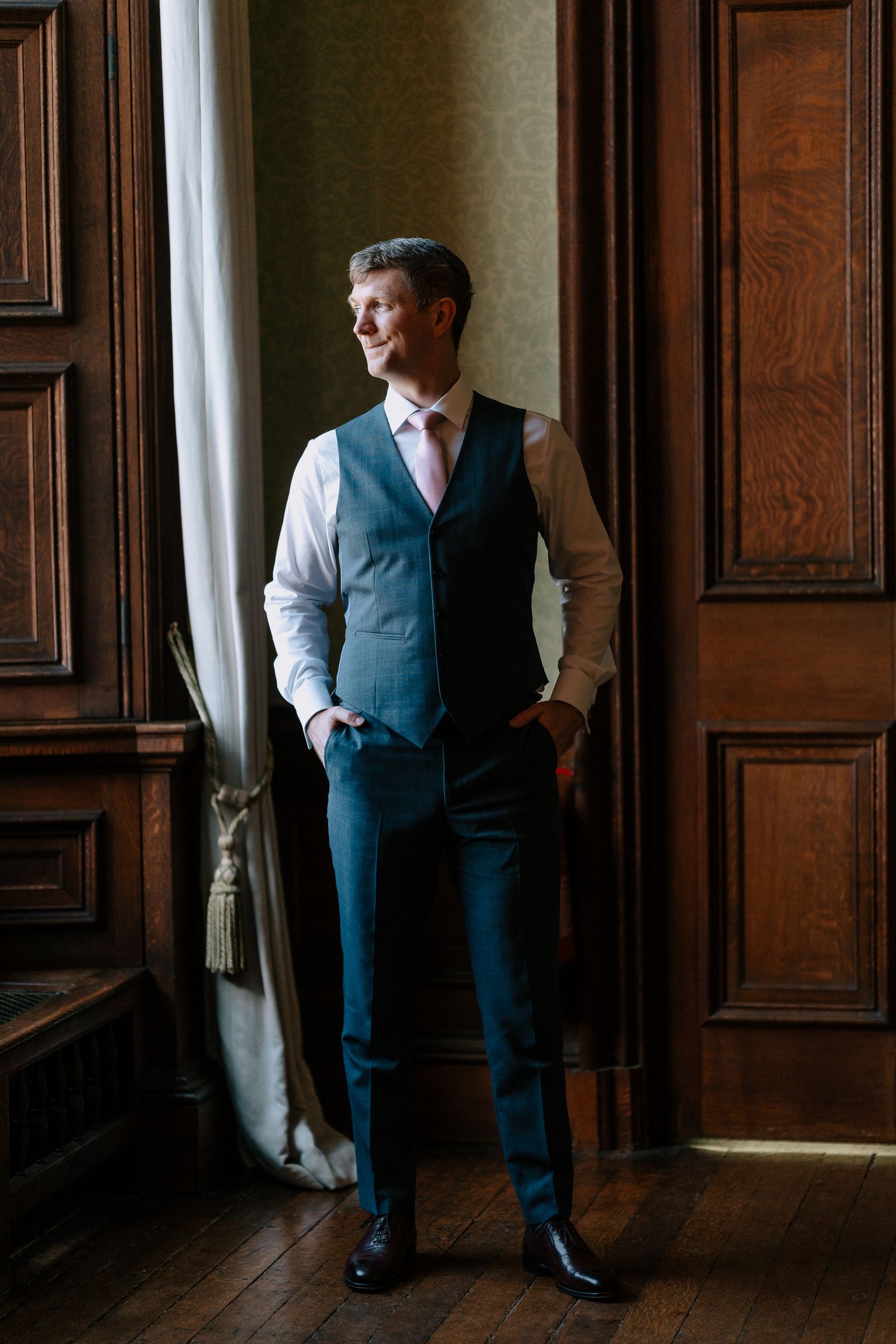 Groom in formal attire poses against wooden paneled walls in classic interior.
