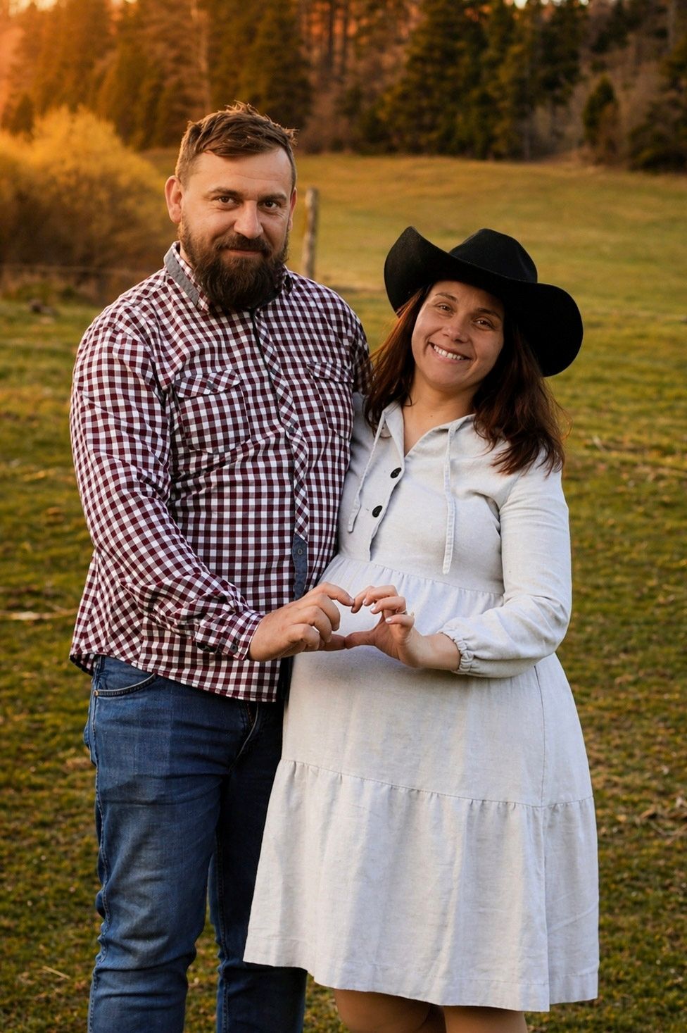 A bearded man in plaid and a woman in a white dress and cowboy hat make a heart shape with their hands outdoors.