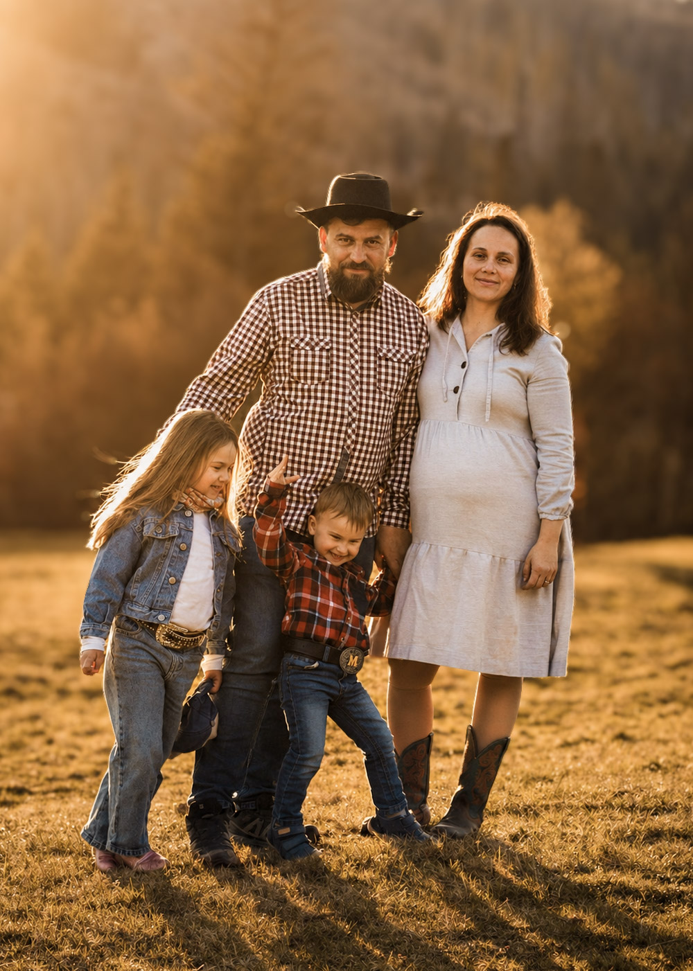 Family of four poses in golden field, pregnant mother in white dress, father in cowboy hat, two young children.