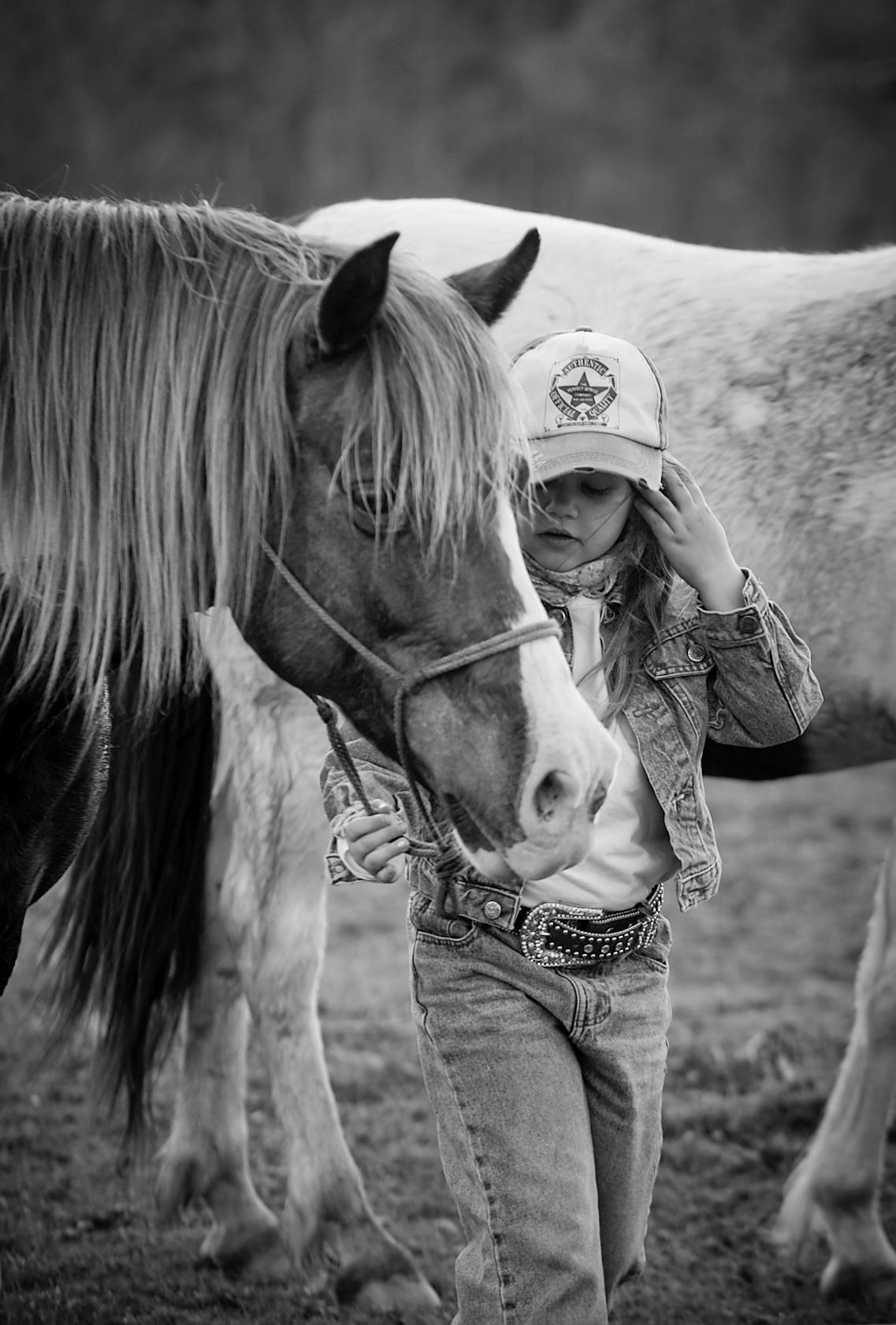 Black and white photo of a young woman in a cowboy hat and denim jacket standing closely with a large horse in a field.