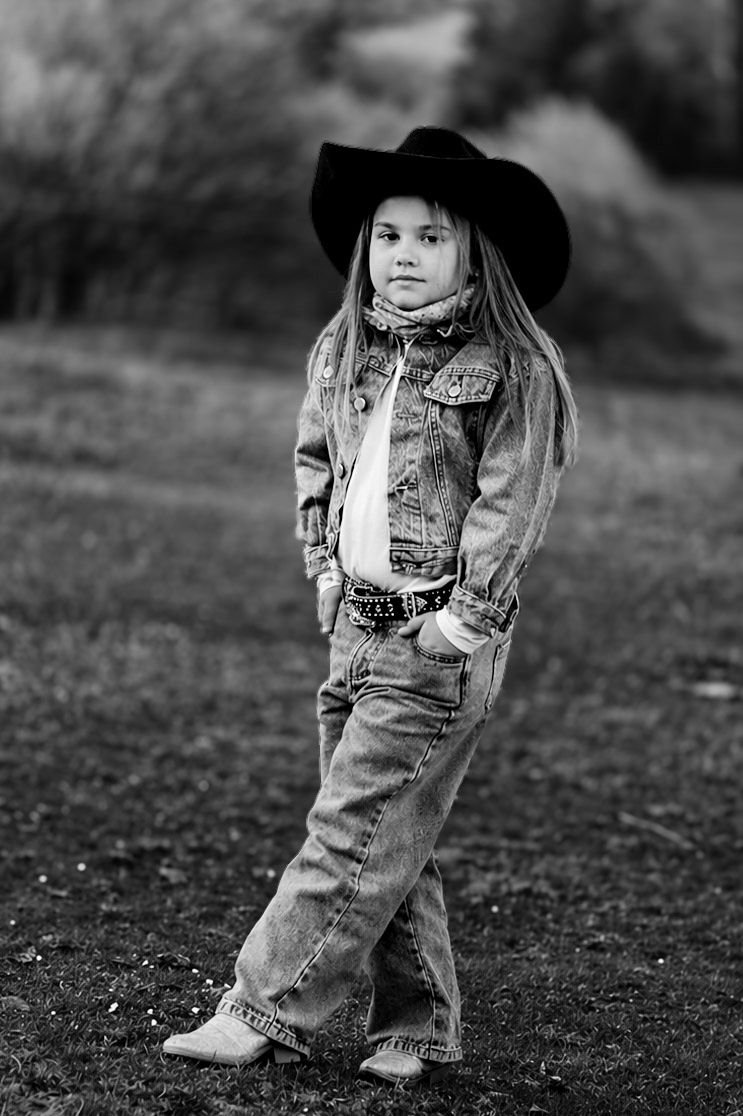 Black and white photo of a young girl in cowboy hat, denim jacket, jeans, and cowboy boots posing outdoors.