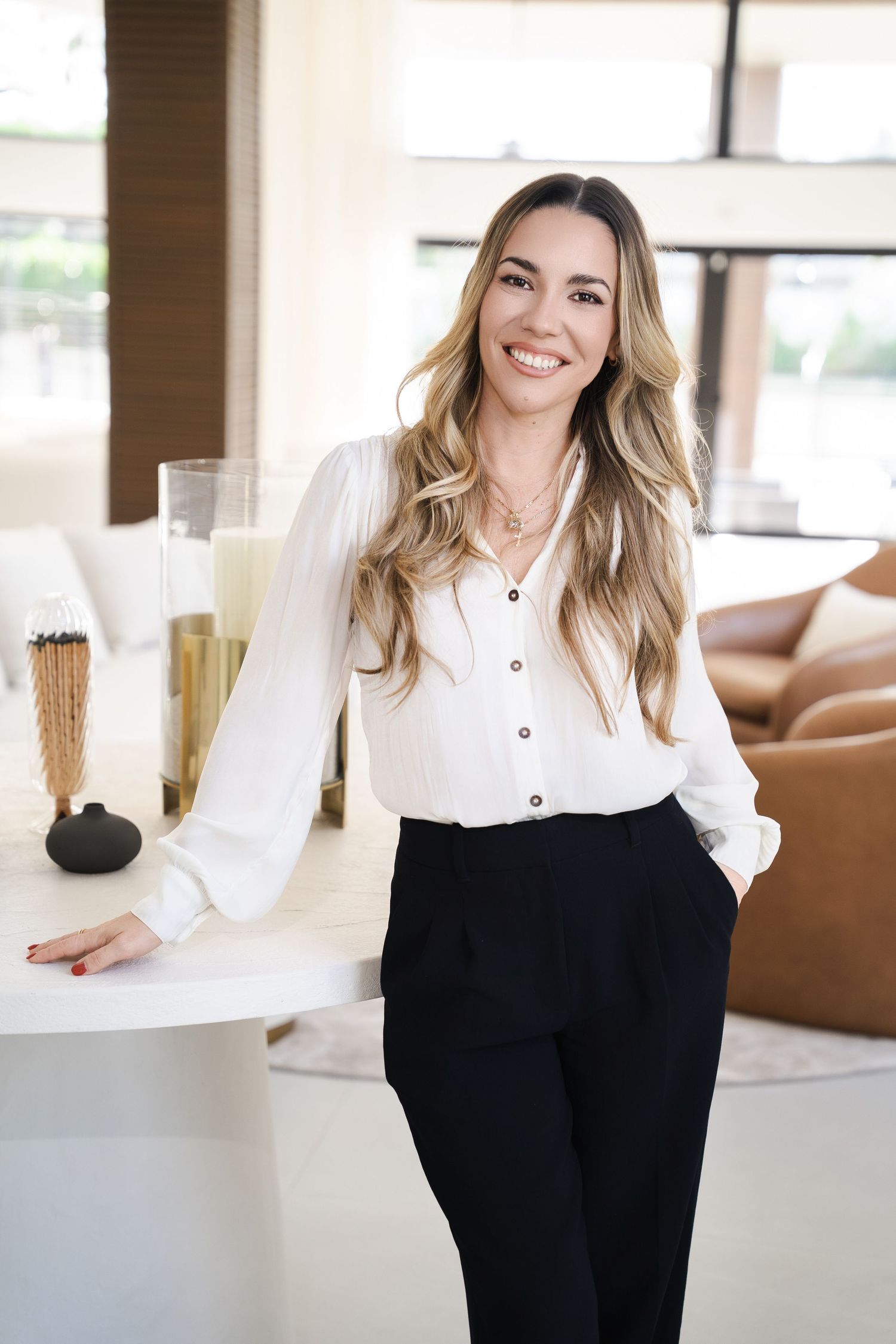 Smiling woman with long blonde hair wearing white blouse and black pants leaning against a white table in a modern interior.