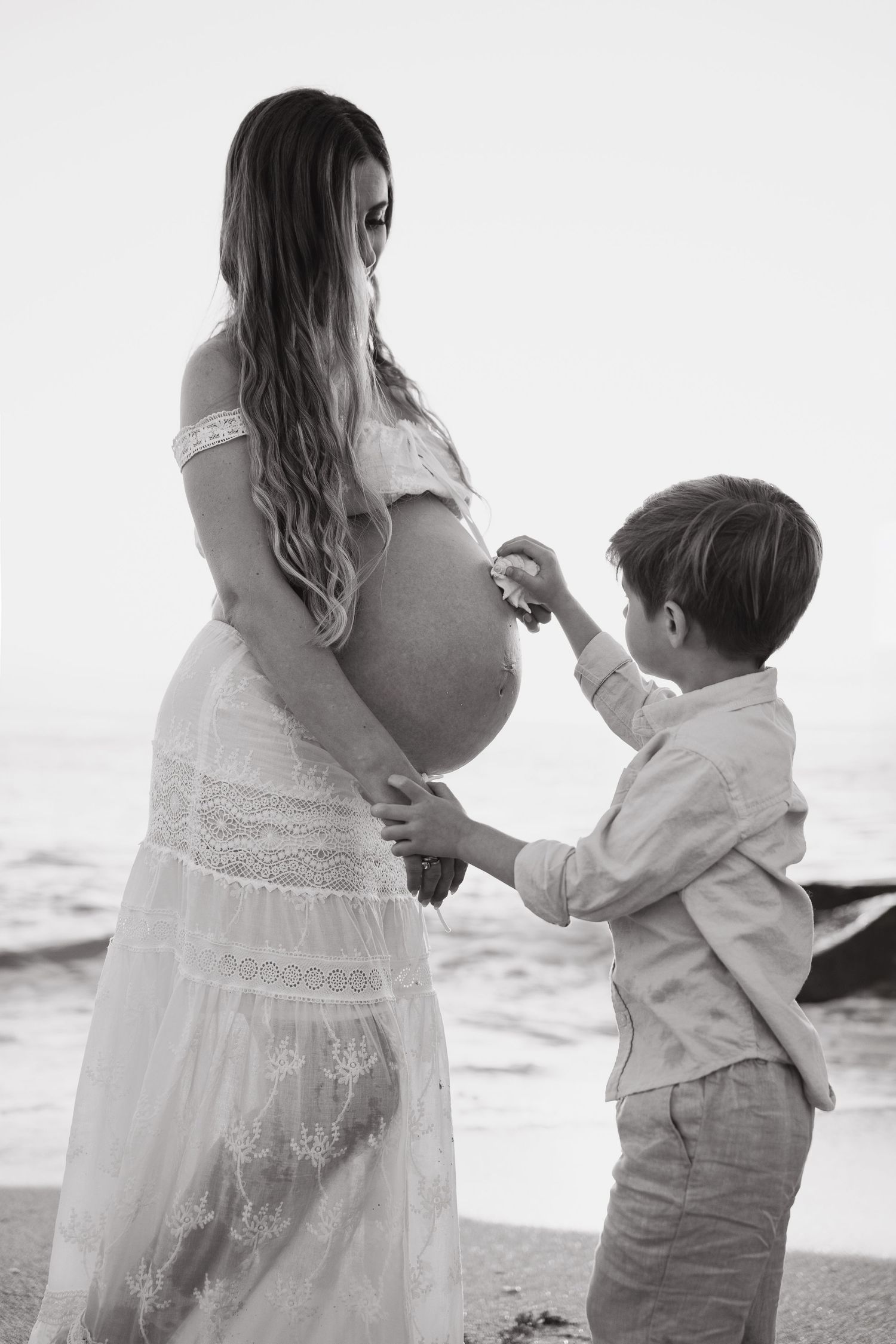 A mother and young son share a tender moment dancing together on a beach in white clothing.