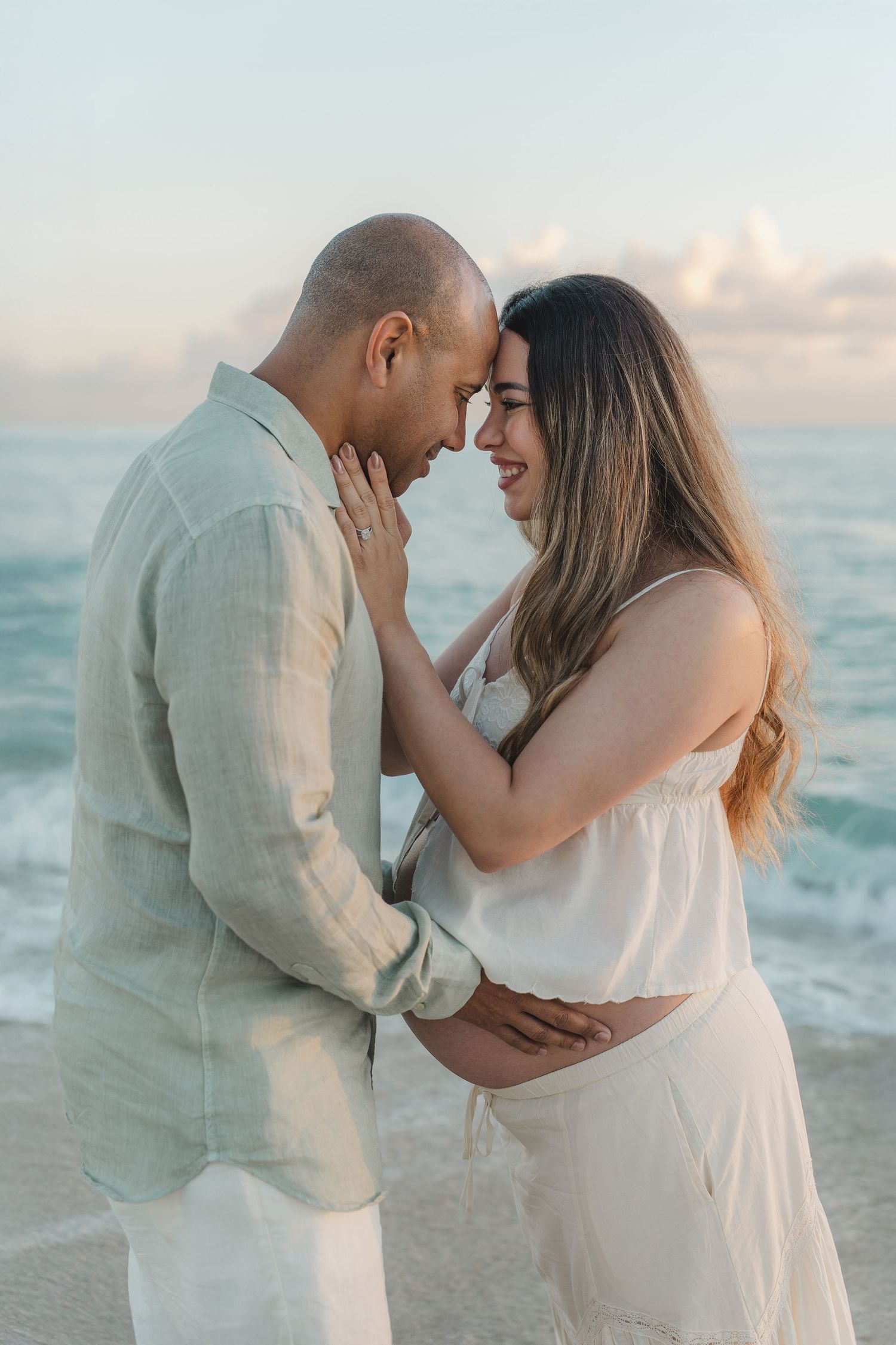 Couple sharing a tender moment on a beach at sunset, woman in white dress with visible baby bump, man in sage linen shirt.