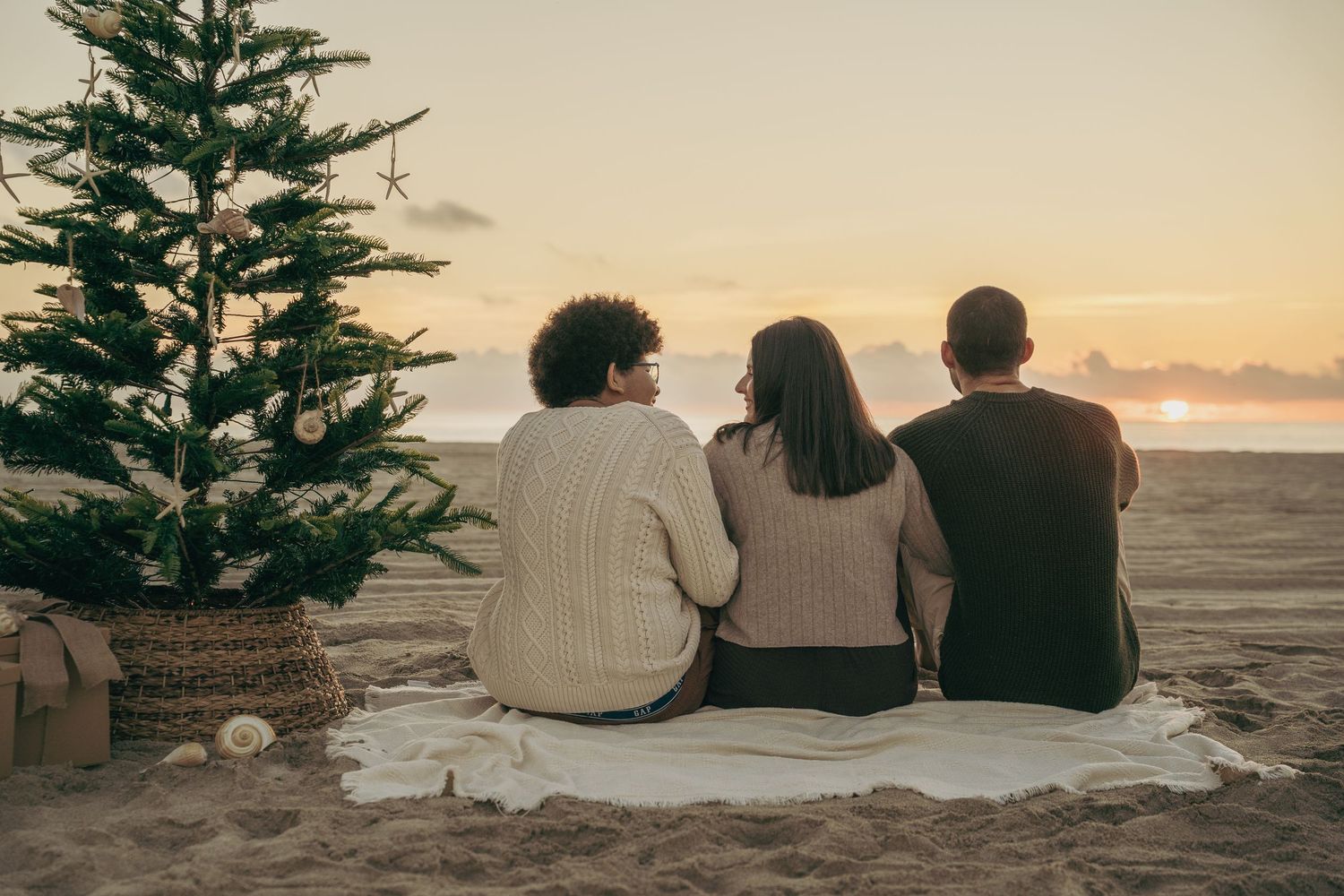 Three people sit on a blanket by a Christmas tree on a sandy beach watching a beautiful sunset together.