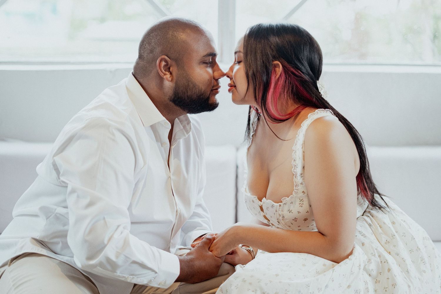 Romantic interracial couple holding hands and sharing a tender moment, nearly kissing, dressed in white outfits indoors.
