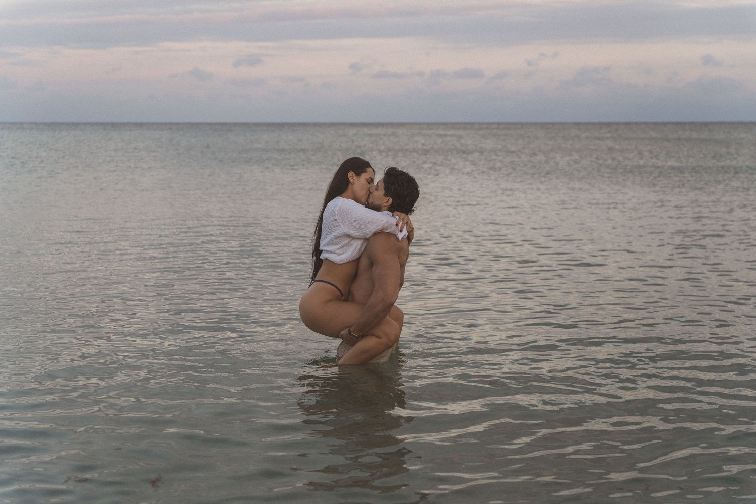 Couple sharing a romantic kiss while standing in shallow ocean water at dusk.