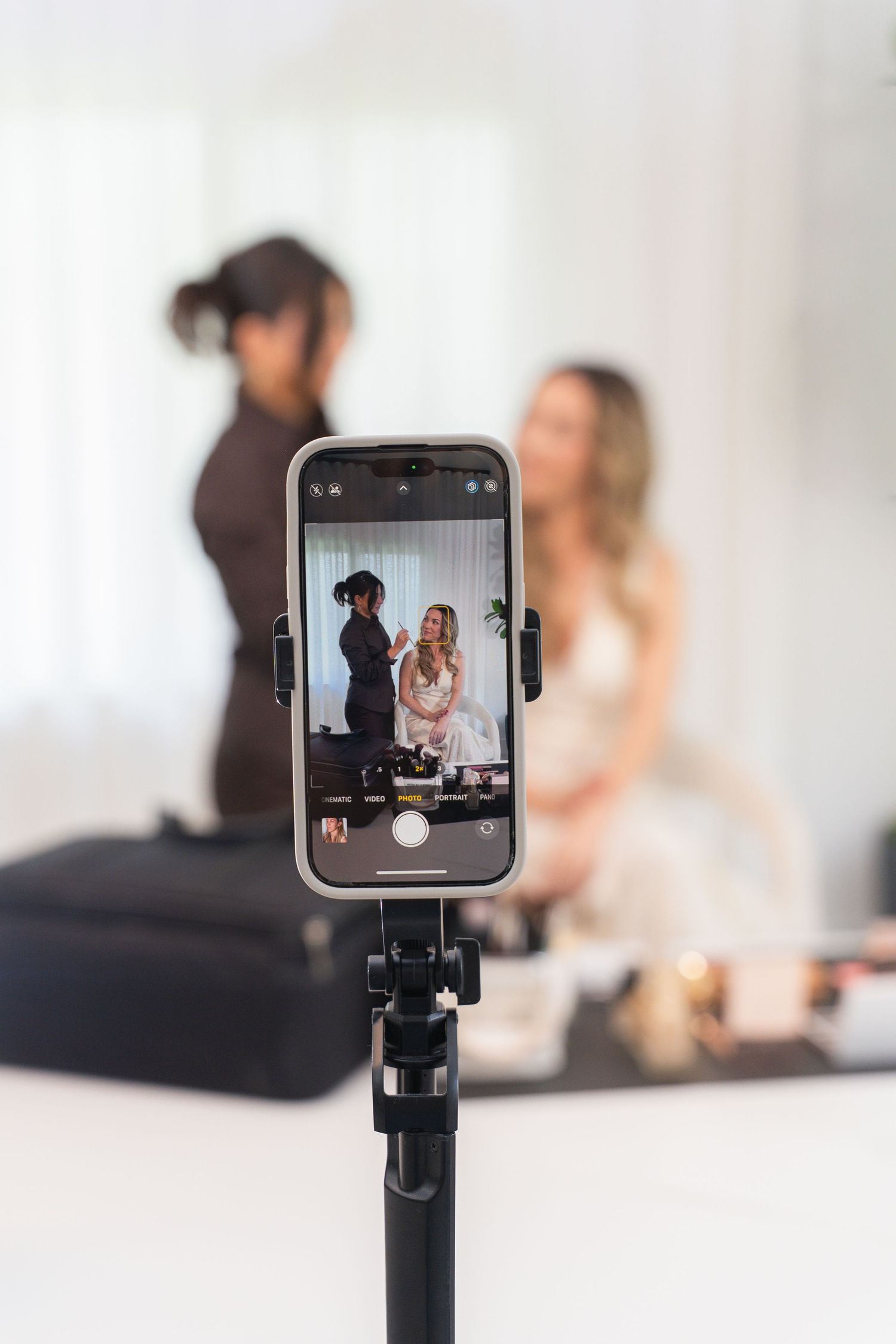 A smartphone on a tripod records a makeup artist applying cosmetics to a bride in a bright studio setting.
