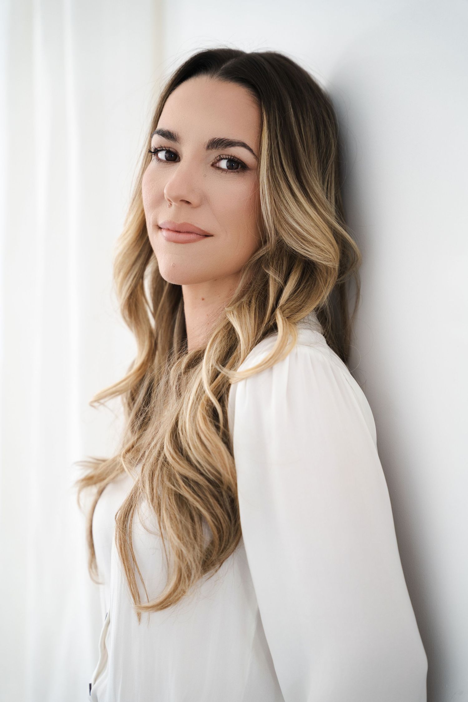 Young woman with long wavy blonde hair wearing a white blouse, posing against a white wall.
