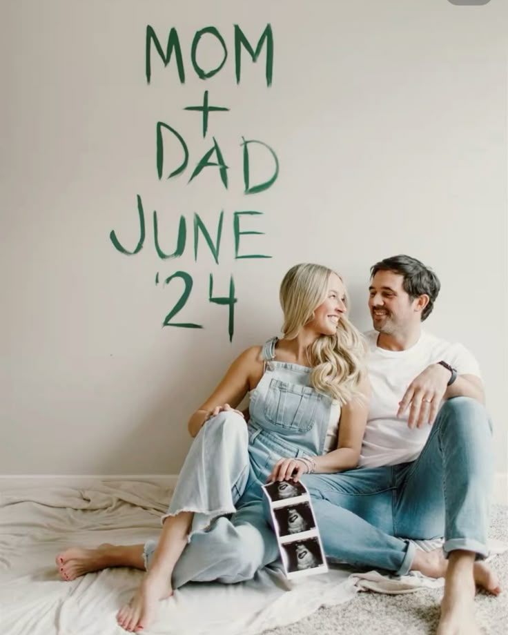 Couple sitting on floor holding ultrasound photos with Mom + Dad June 24 pregnancy announcement on wall.