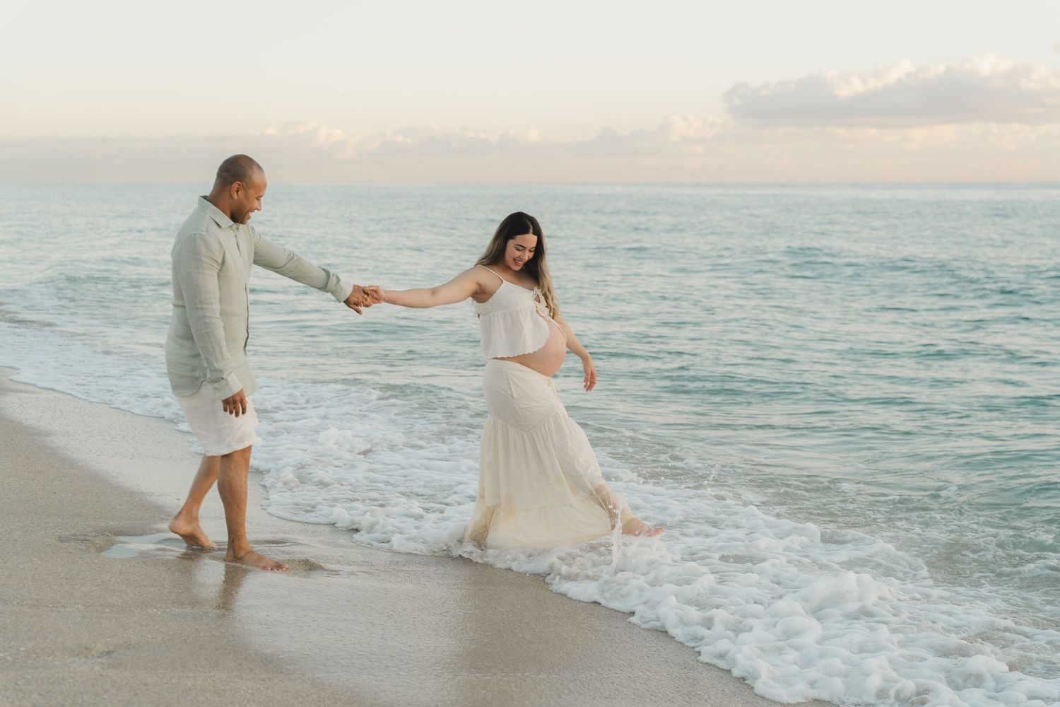 Candid moment of couple playing in ocean waves during maternity beach session.