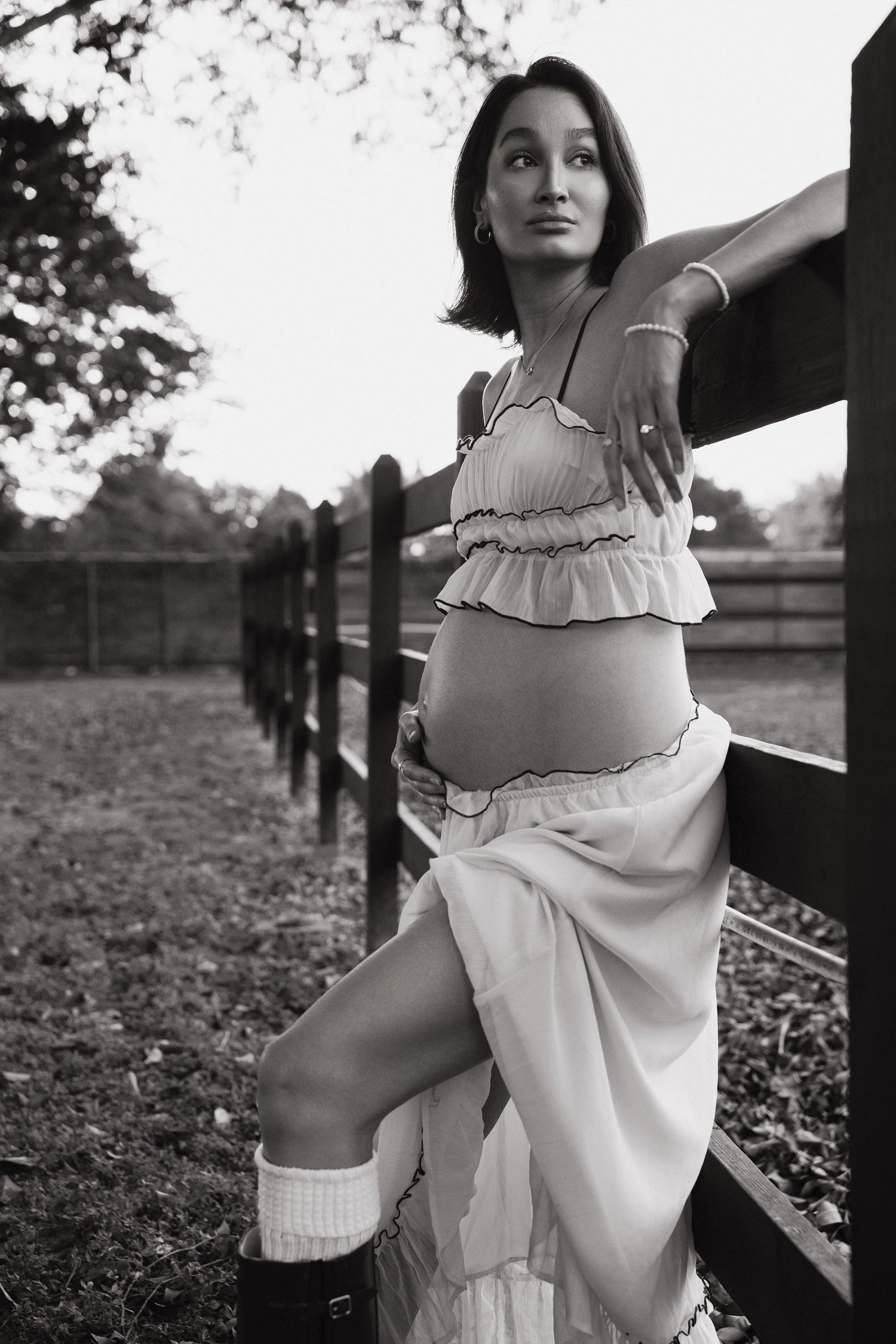 Black and white photo of a pregnant woman in ruffled crop top and skirt leaning against a wooden fence outdoors.