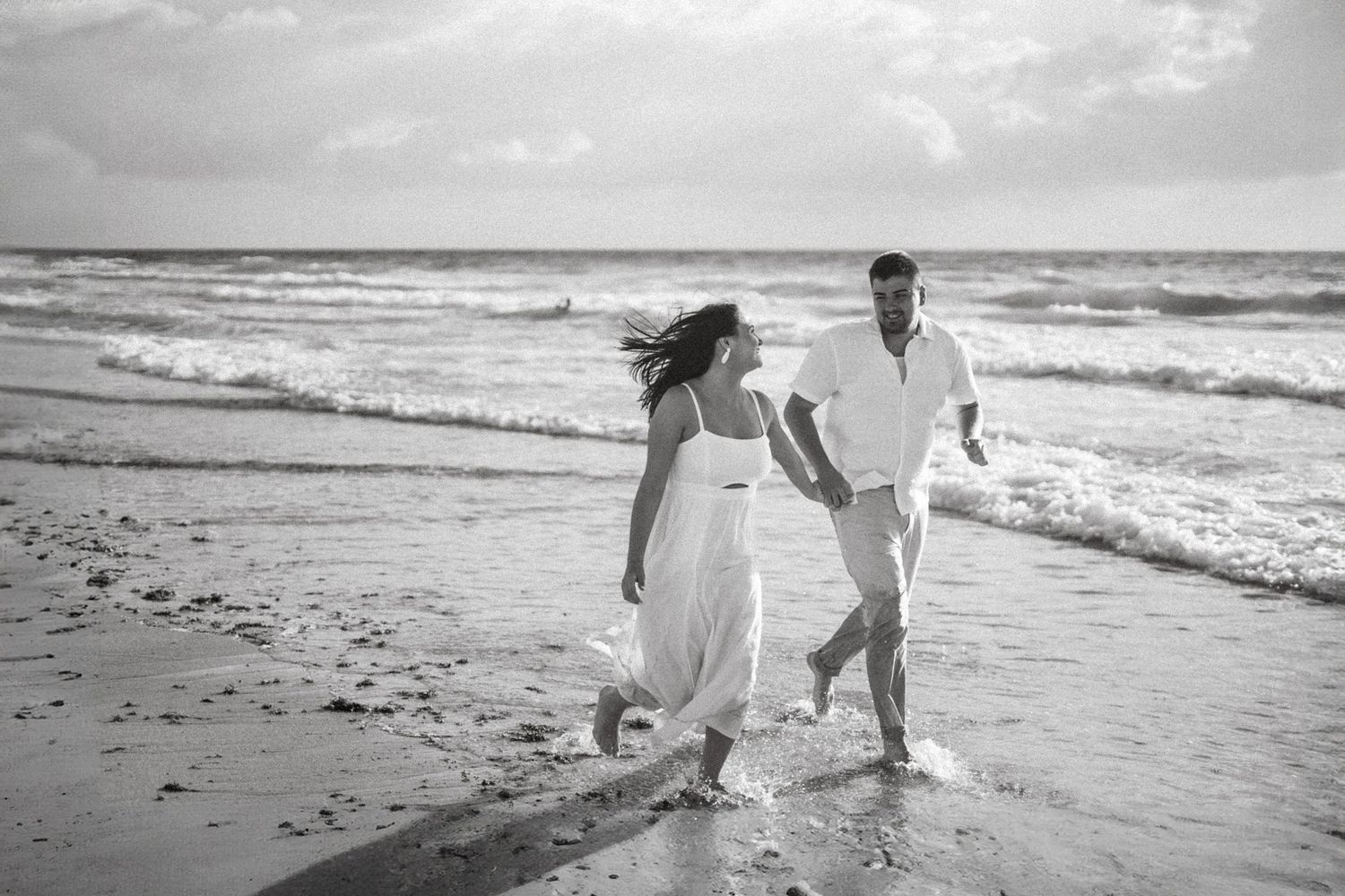 Black and white photograph of a couple running joyfully along the beach shoreline.