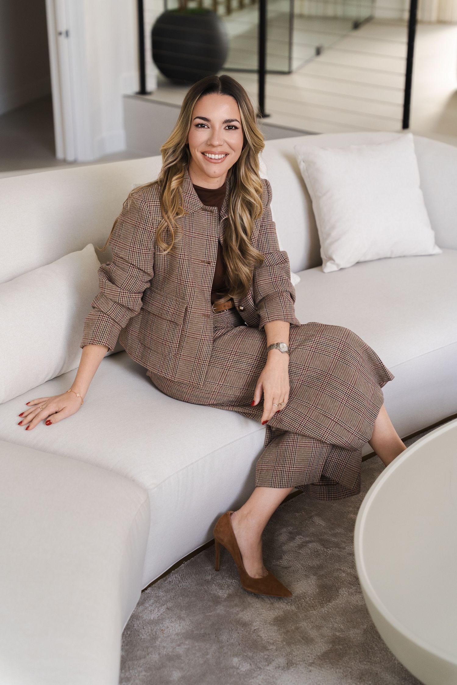 Woman in brown plaid suit sitting elegantly on a white curved sofa in a modern living room.