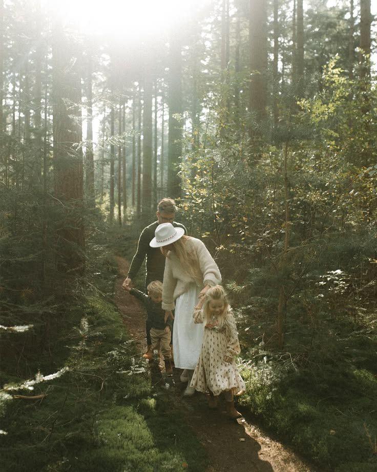 Family walking on a sunlit forest path, adult in white hat and dress guiding two young children through tall trees.
