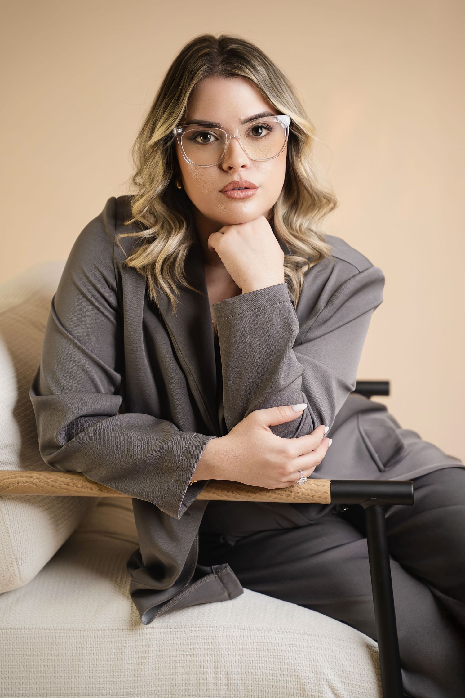Woman wearing gray blazer and glasses poses on beige couch during indoor lifestyle photoshoot.