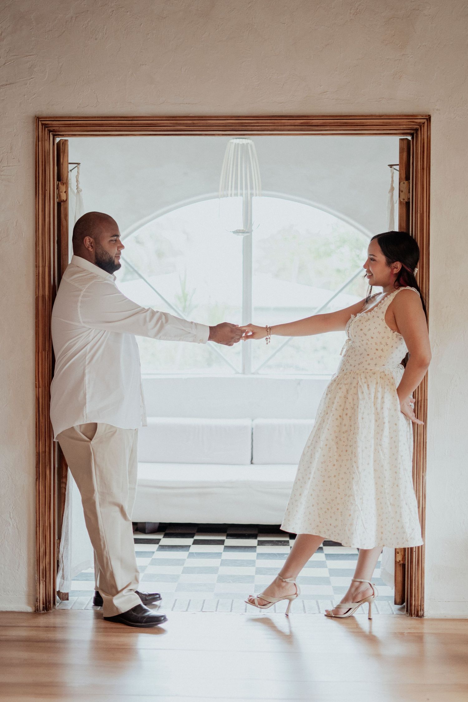 A couple in white outfits dances playfully in a bright minimalist room with wooden window frames.