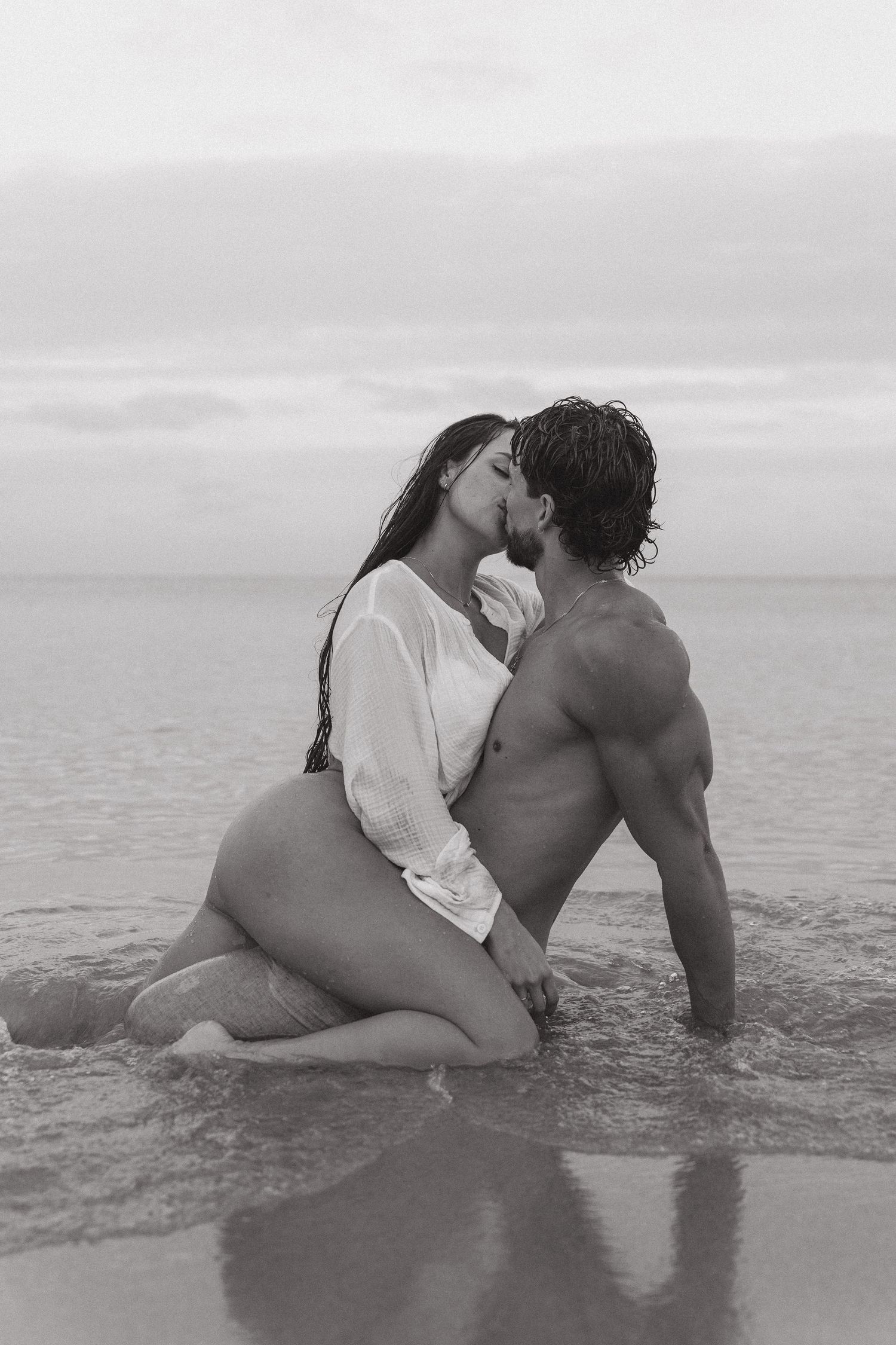 Black and white photo of a romantic couple kissing on the shoreline, sitting in shallow ocean waves.