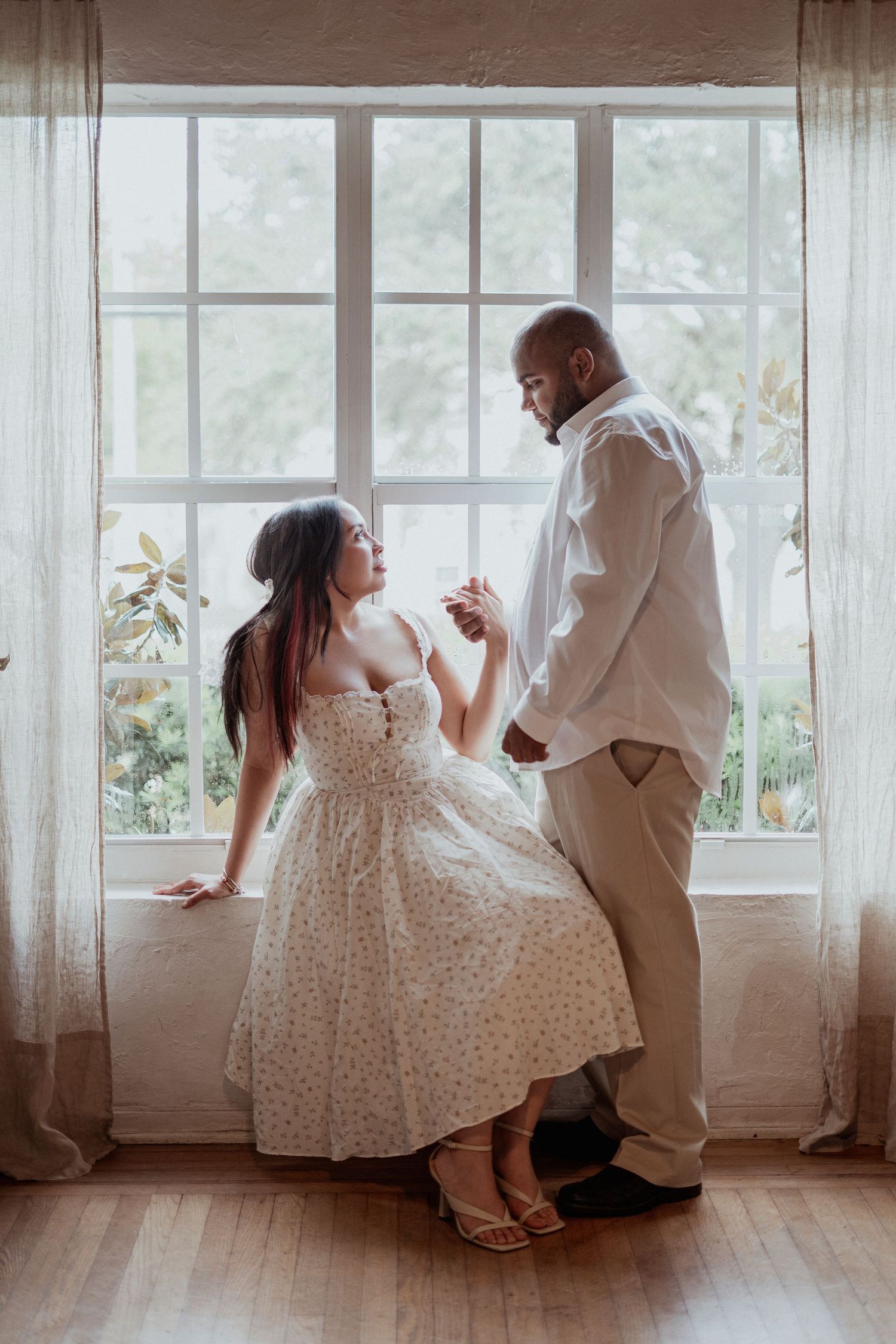Couple sharing a romantic moment by a large window, woman in floral dress and man in white shirt holding hands indoors.