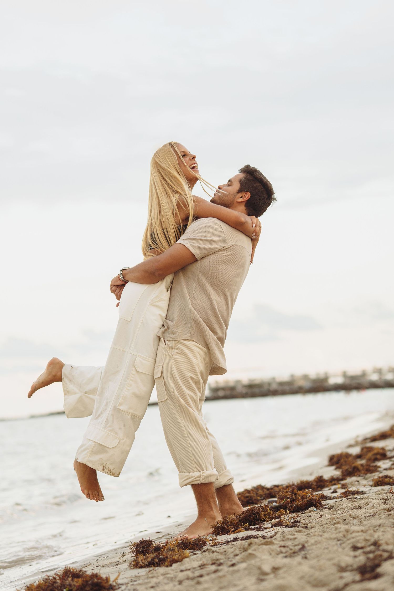 A couple in beige outfits embraces playfully on the beach.
