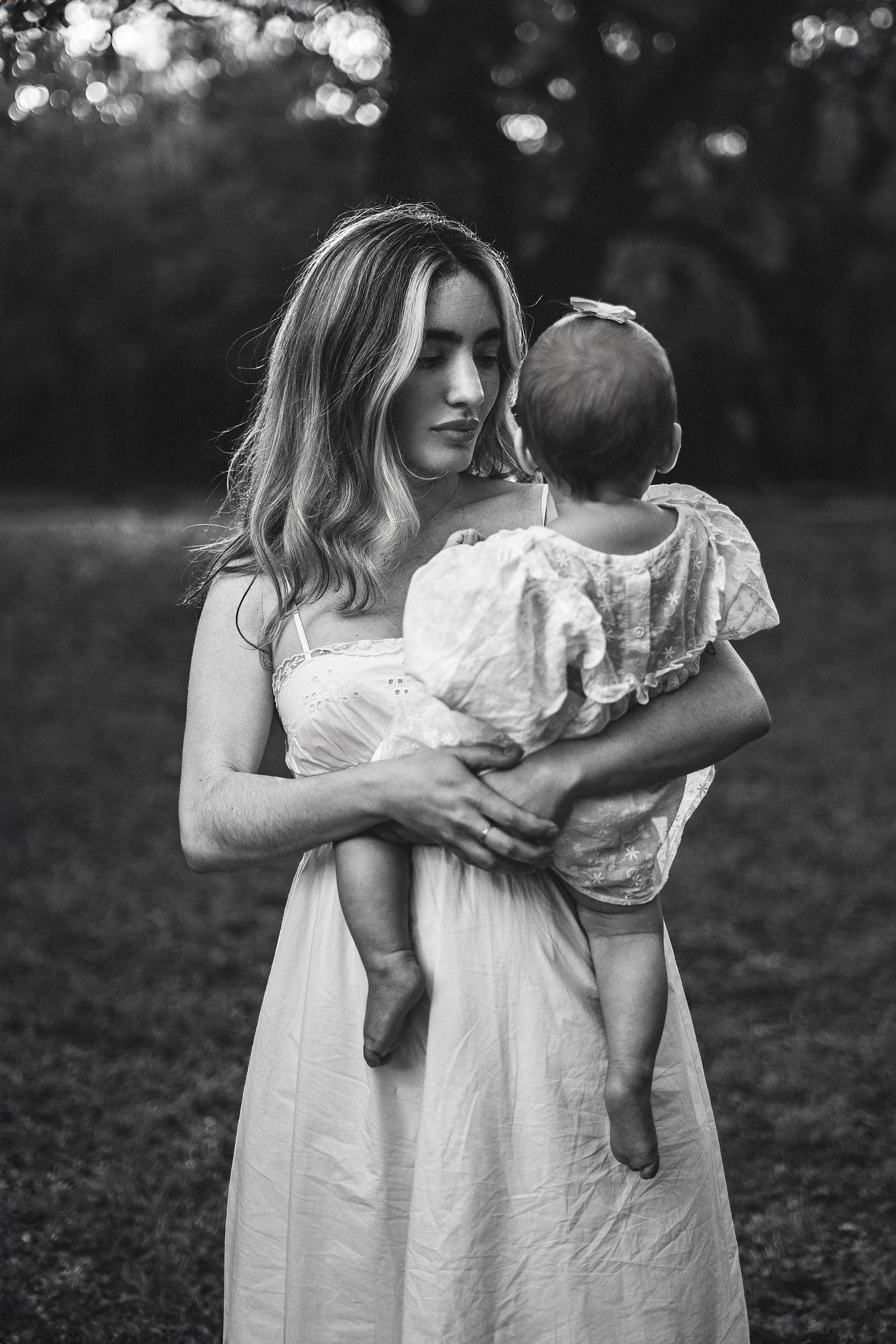 Black and white portrait of mother holding baby in white dress outdoors.