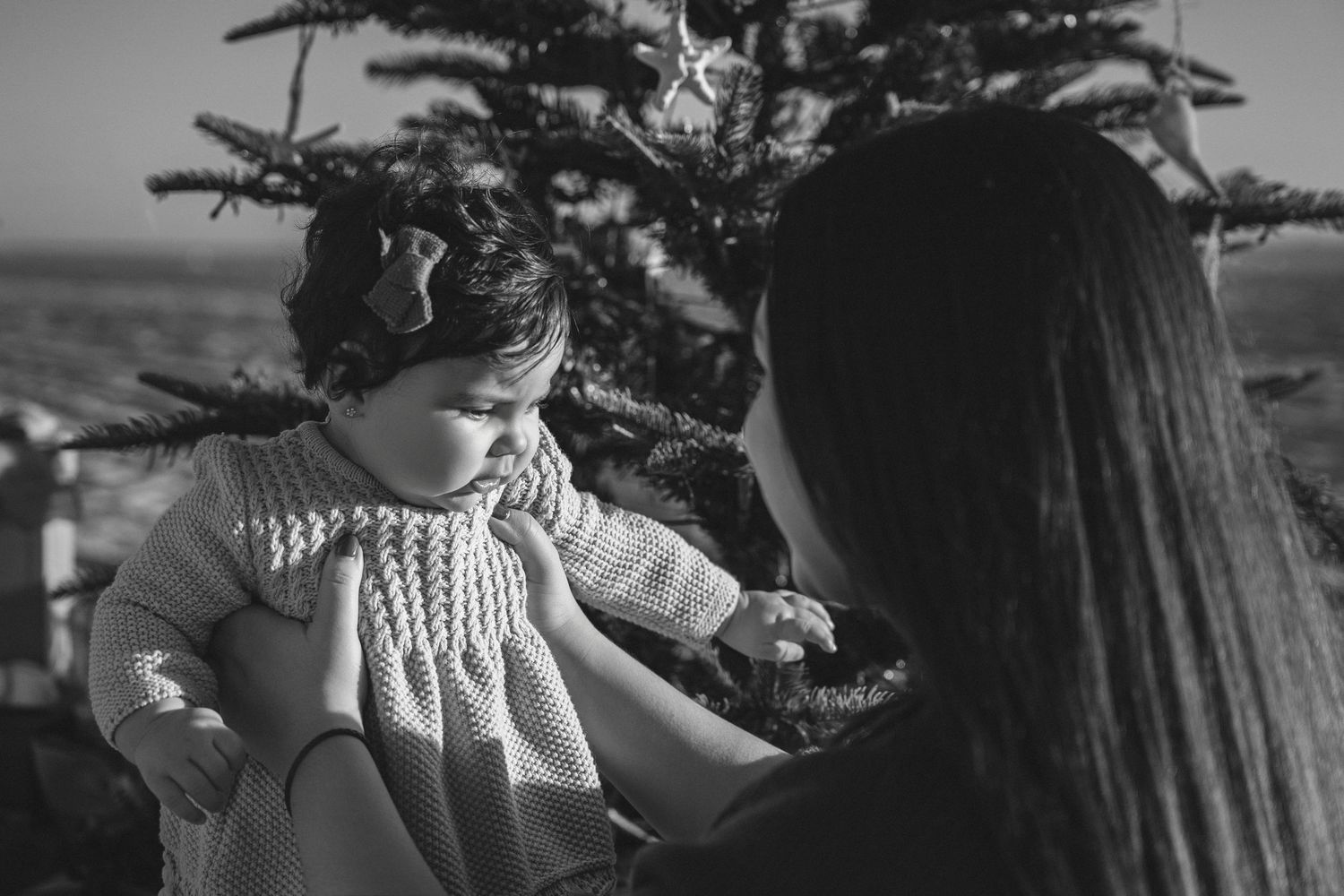 Black and white photo of a mother holding her baby girl near a Christmas tree outdoors.