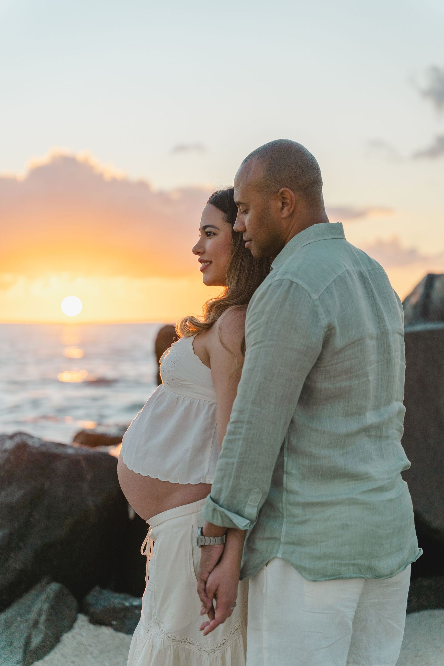 Expecting couple shares intimate moment at sunset on rocky beach.