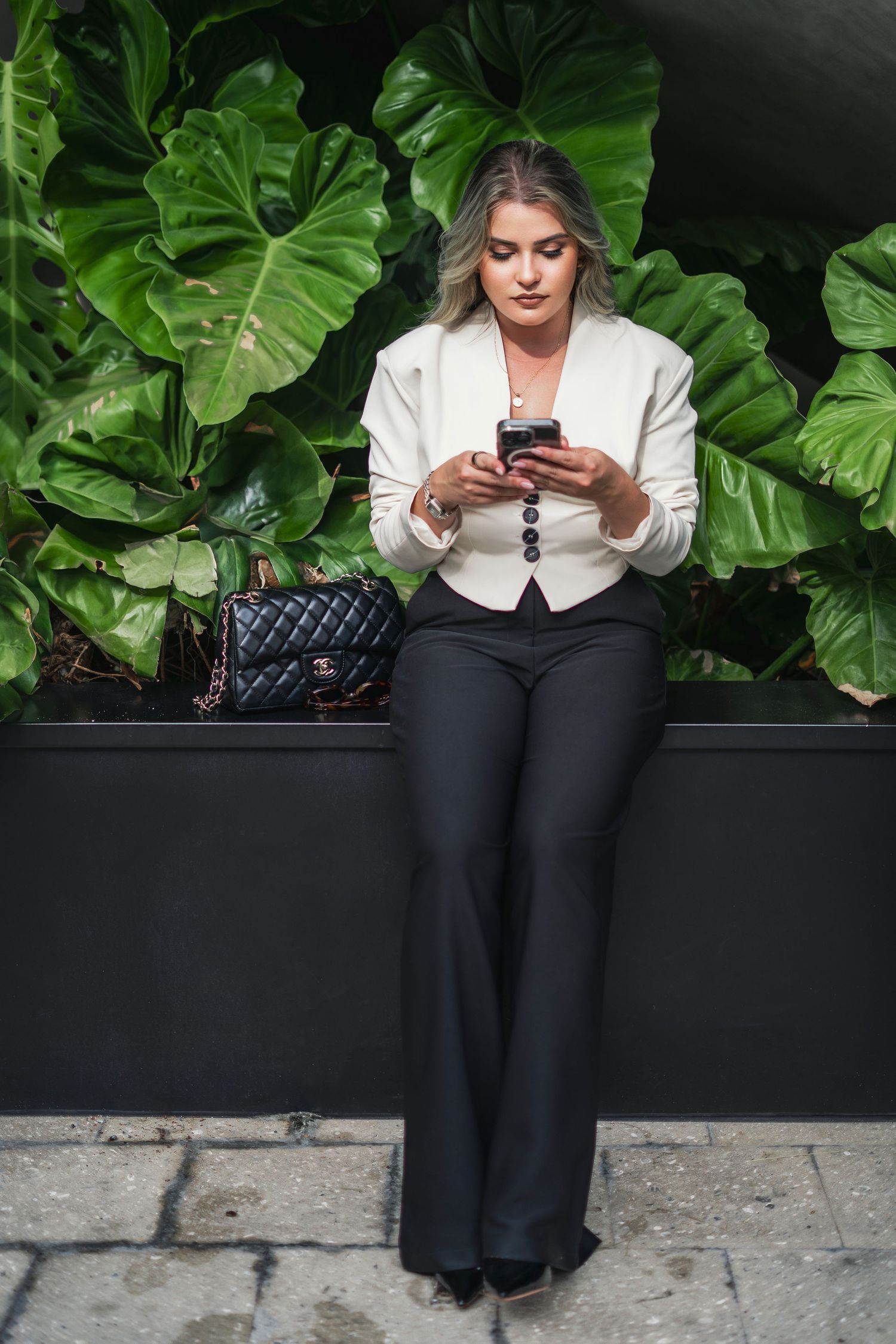 Person in white blazer and black pants sitting with phone against tropical plant backdrop.