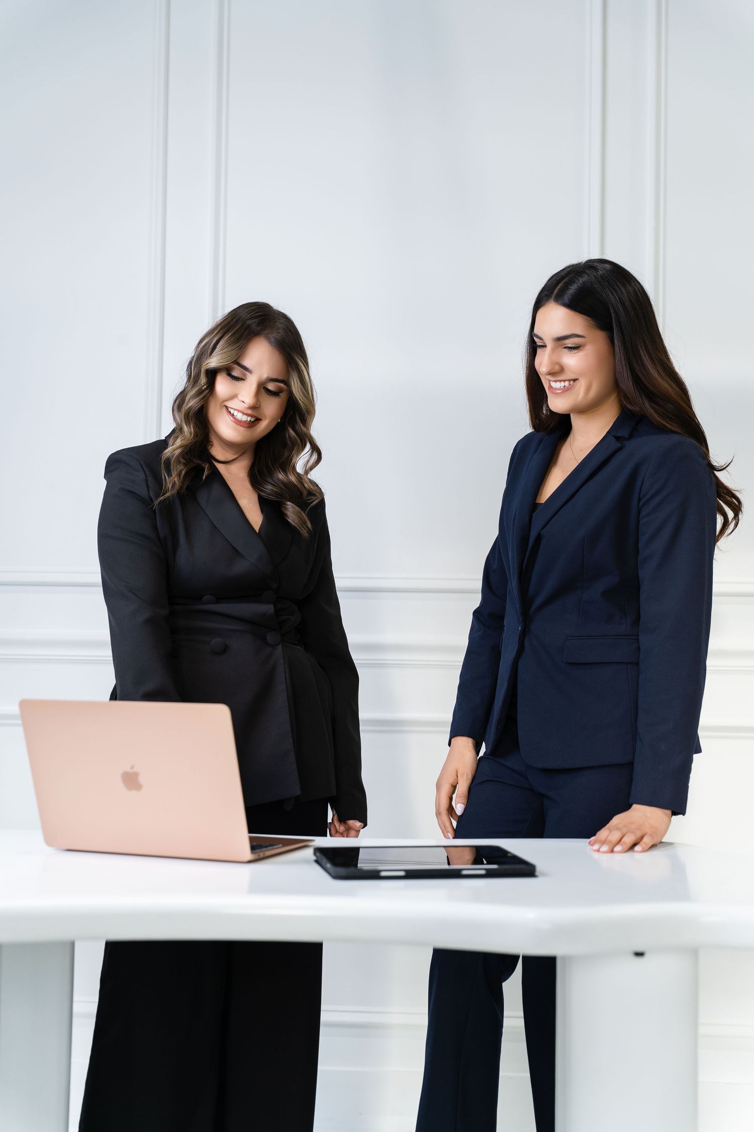 Two professional women in suits smiling while working together at a white desk with a laptop and tablet.