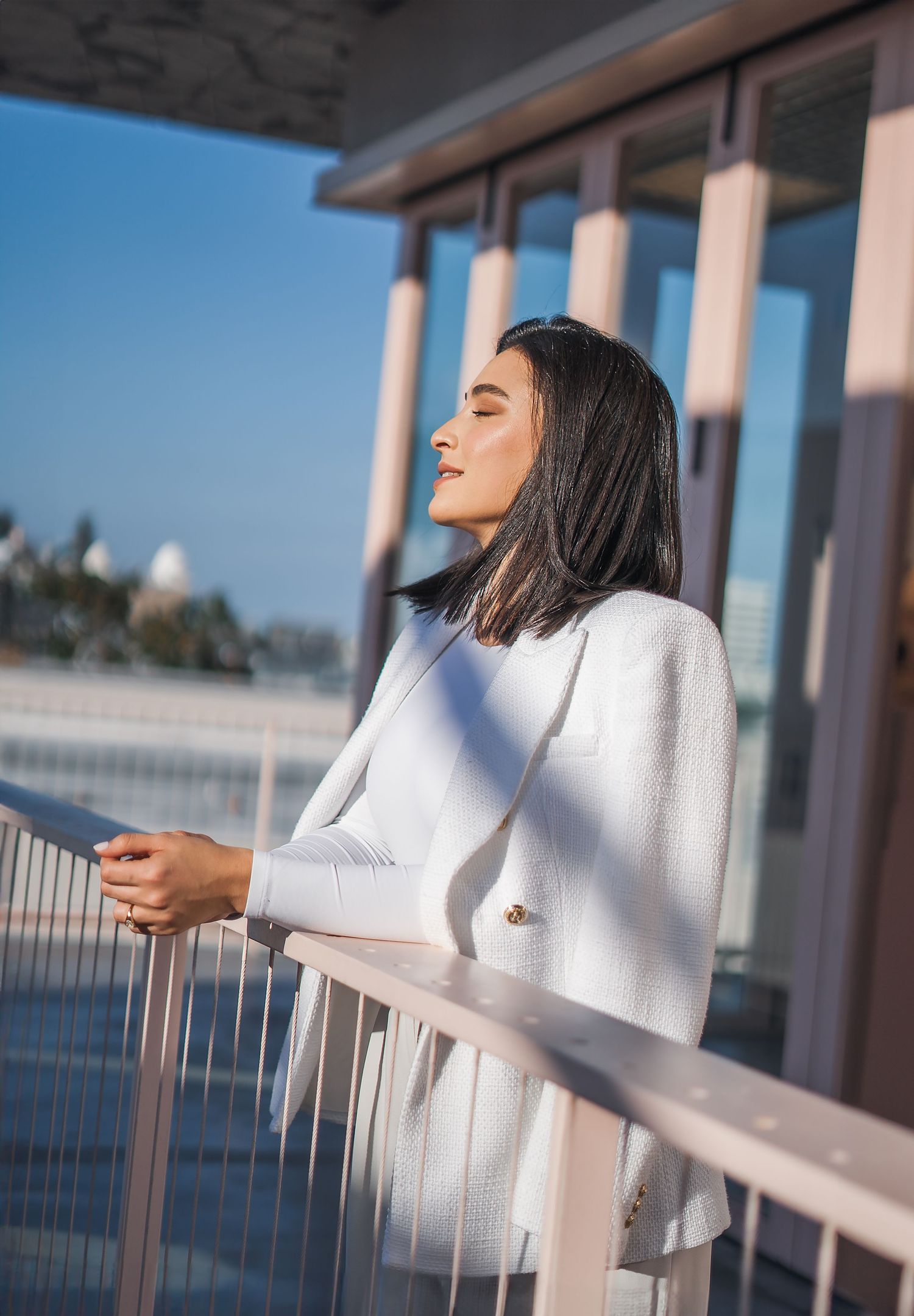 Woman in white sweater smiles while leaning on balcony railing during outdoor photoshoot.