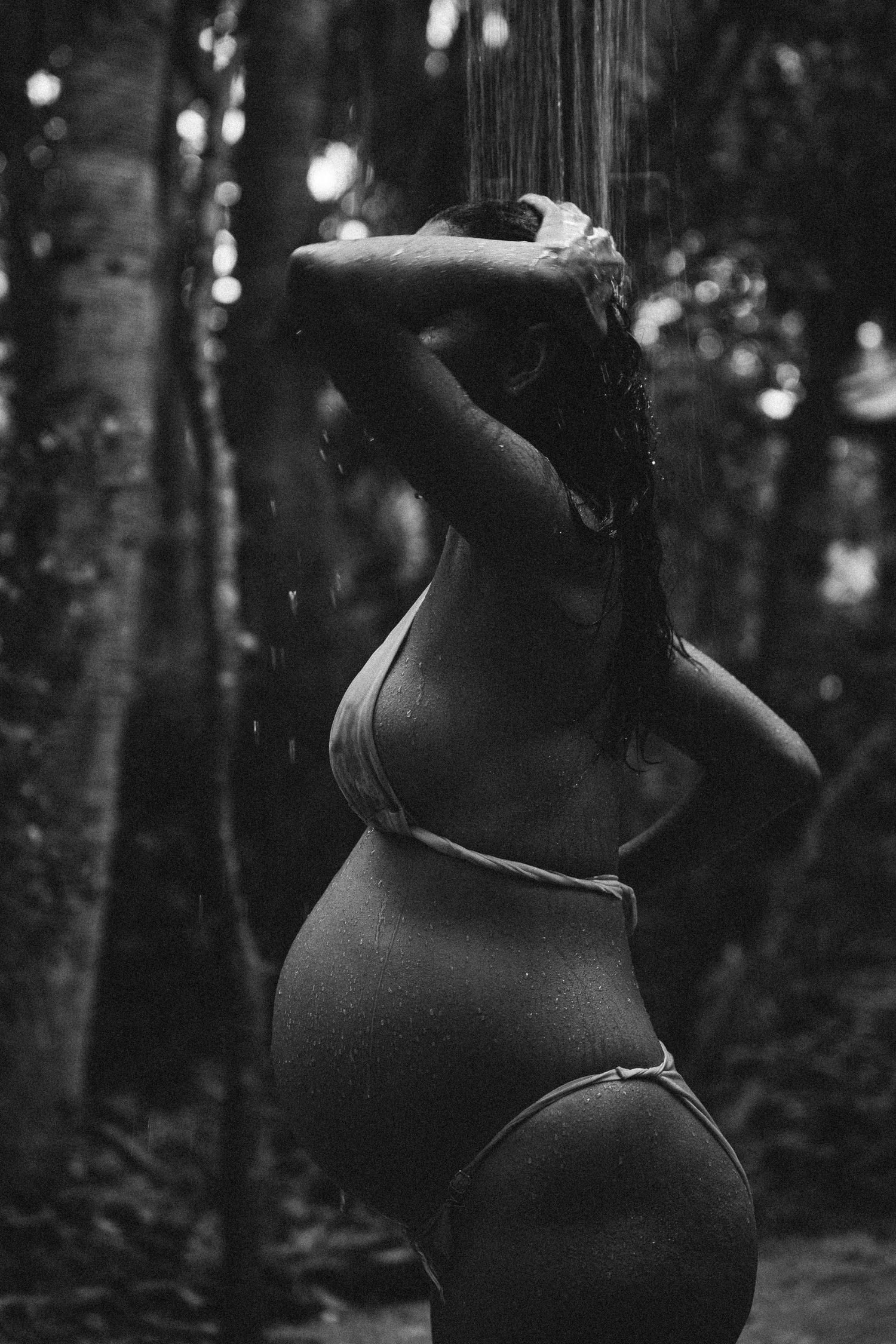 Black and white photo of a pregnant woman showering outdoors in a forest setting.