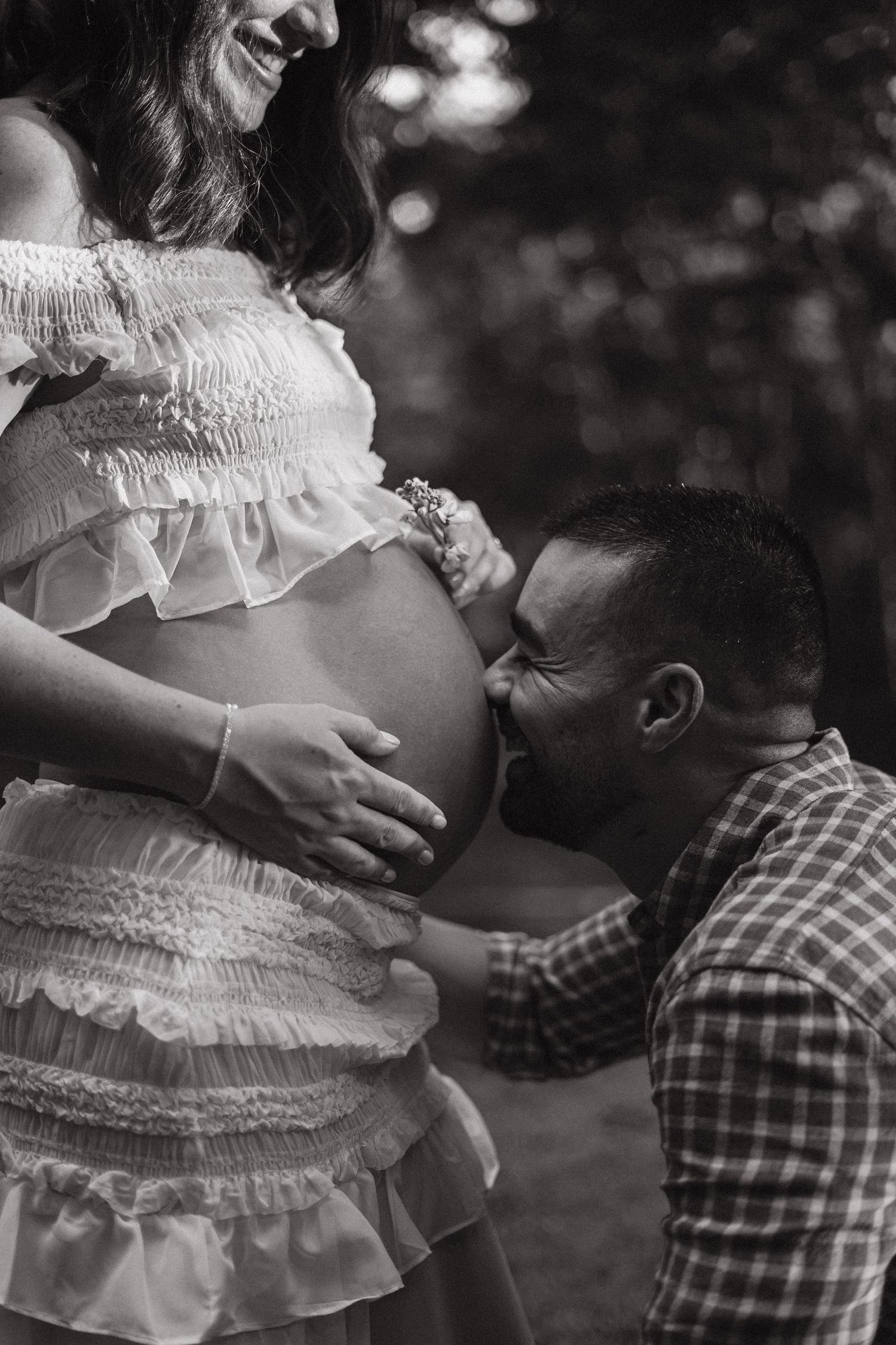 Black and white maternity photo of a man kissing pregnant woman's belly outdoors.