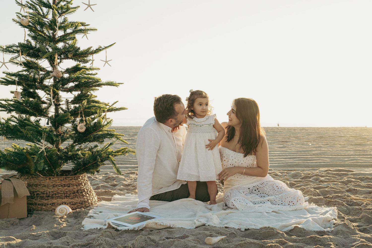 Family of three sitting on beach blanket by decorated Christmas tree, parents kissing toddler in white dress at sunset.