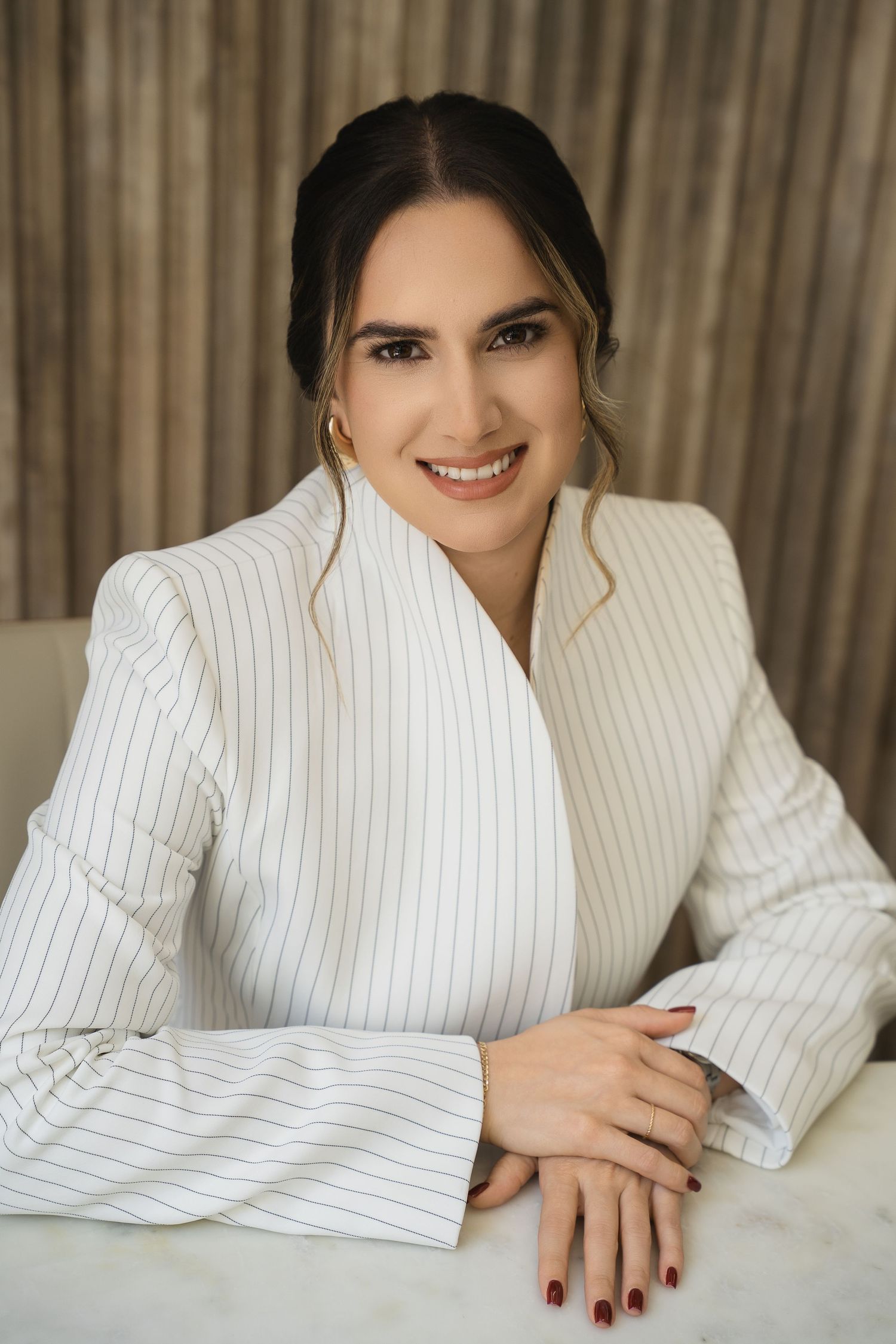 Smiling woman in white pinstripe blazer seated at table, dark hair up, red nails, hoop earrings, professional portrait.