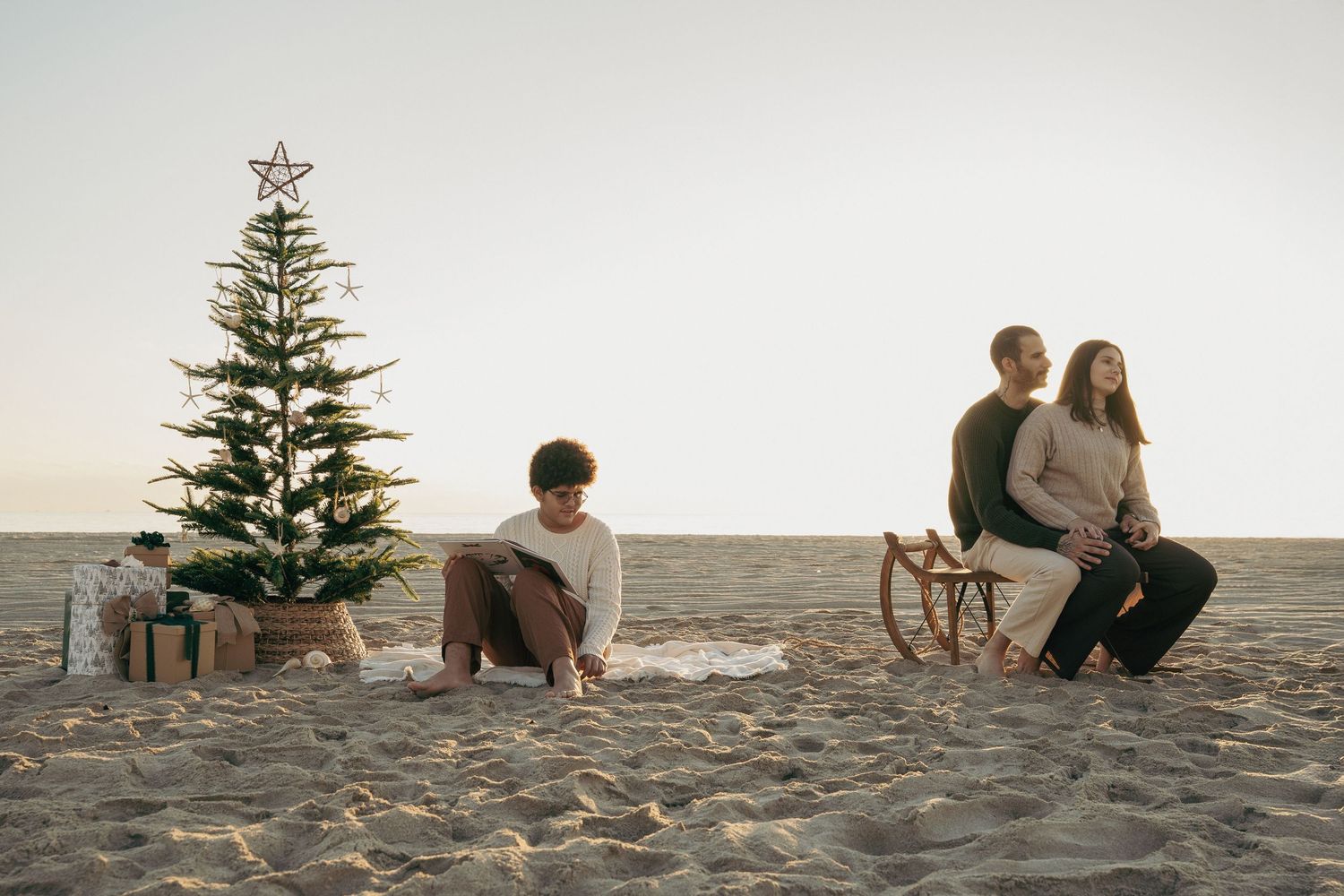 Family celebrating Christmas on a sandy beach with a decorated tree, gifts, and a wooden sled at sunset.
