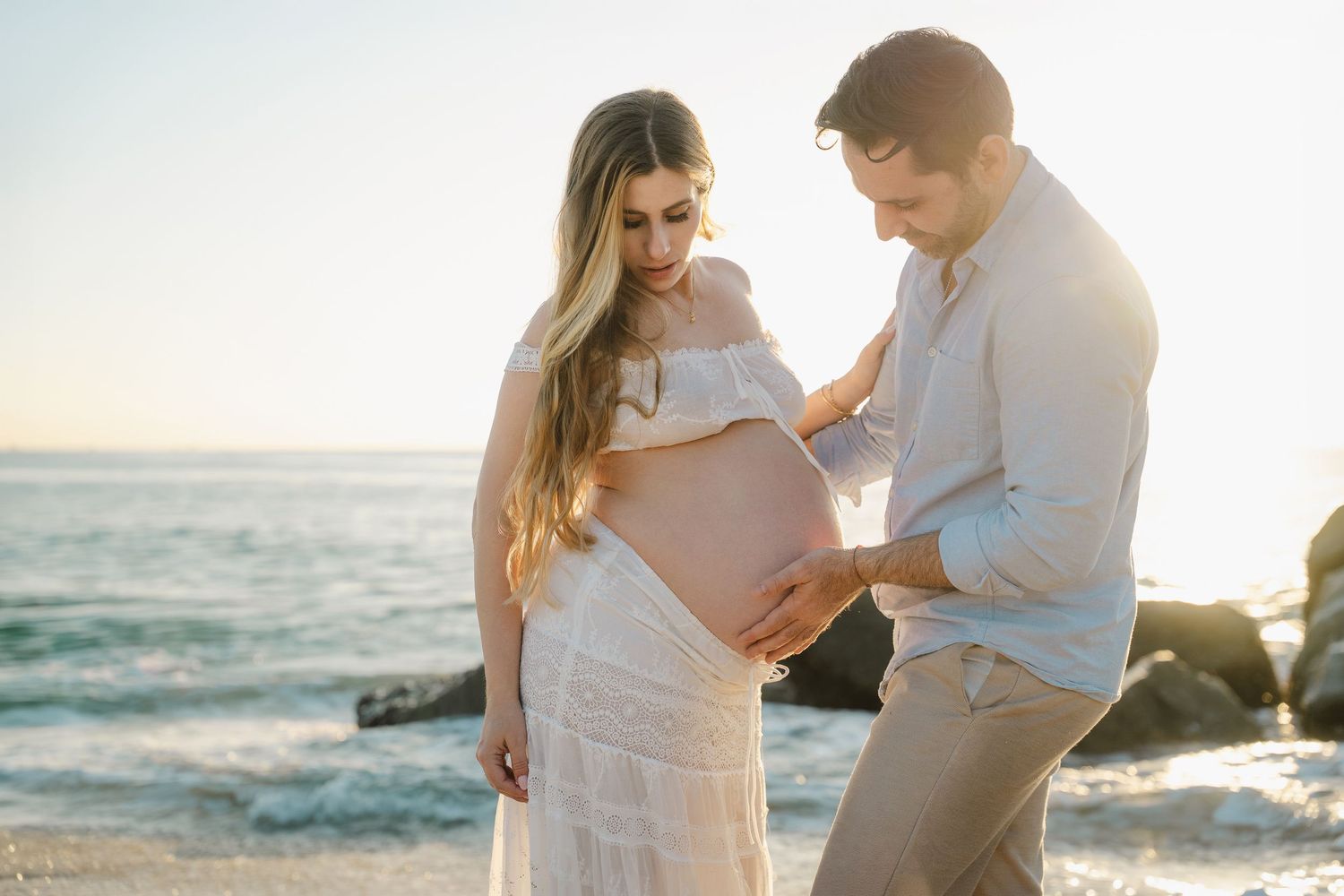 A couple shares an intimate moment on a sunlit beach as they celebrate an upcoming arrival at sunset.