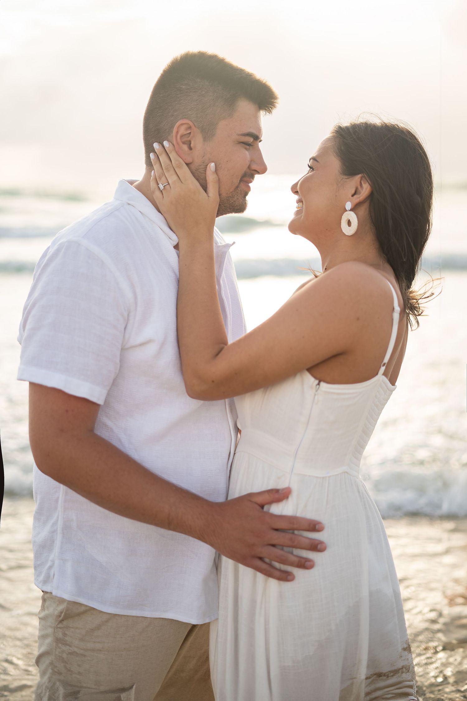 A romantic couple embraces on the beach at sunset wearing white and beige outfits.