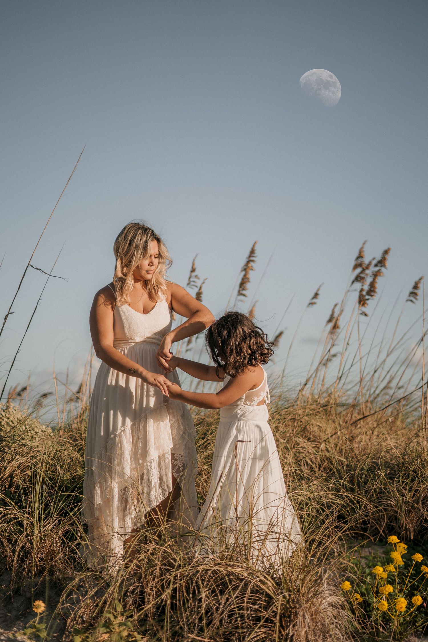 Mother and daughter in white dresses share playful moment among coastal grasses at dusk.