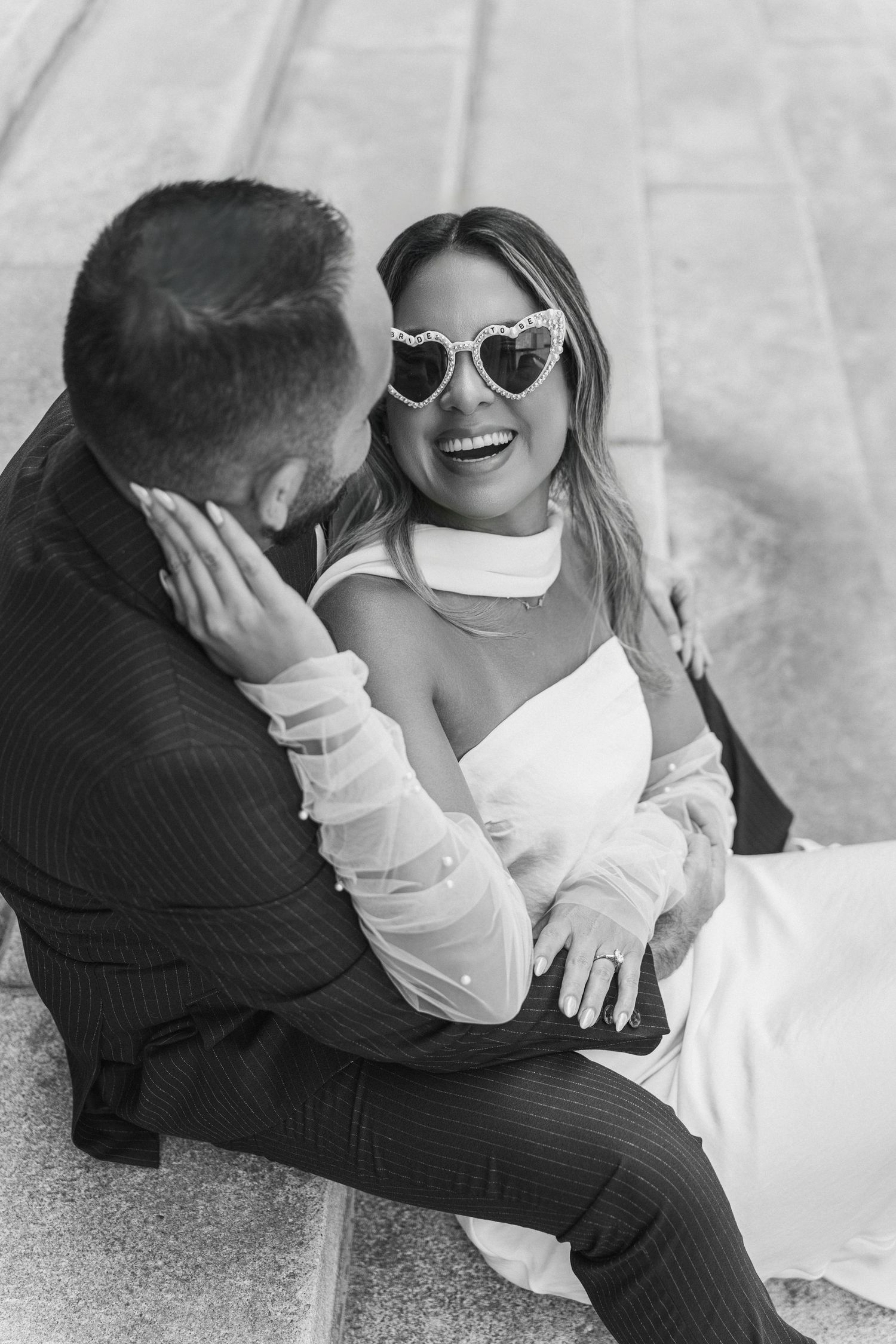 A playful couple in white wedding attire laughs together while wearing sunglasses outdoors.