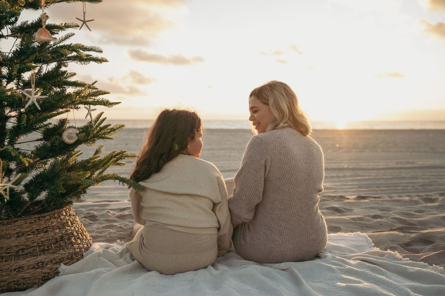 Two women sit on a beach blanket by a Christmas tree watching a golden sunset together.