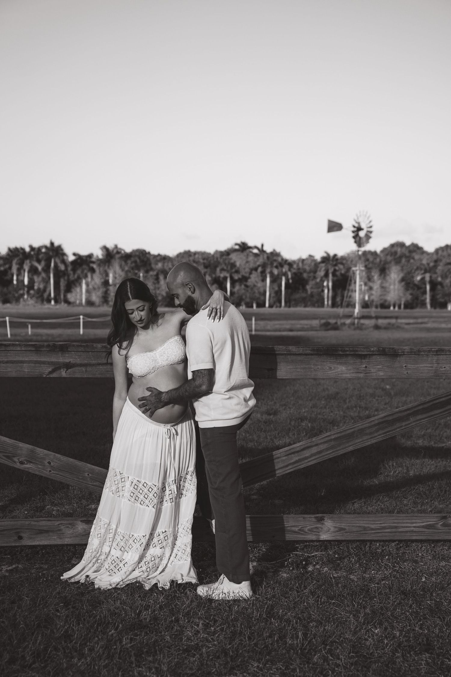 Black and white photo of a couple embracing outdoors near a wooden fence, with palm trees and a windmill in the background.