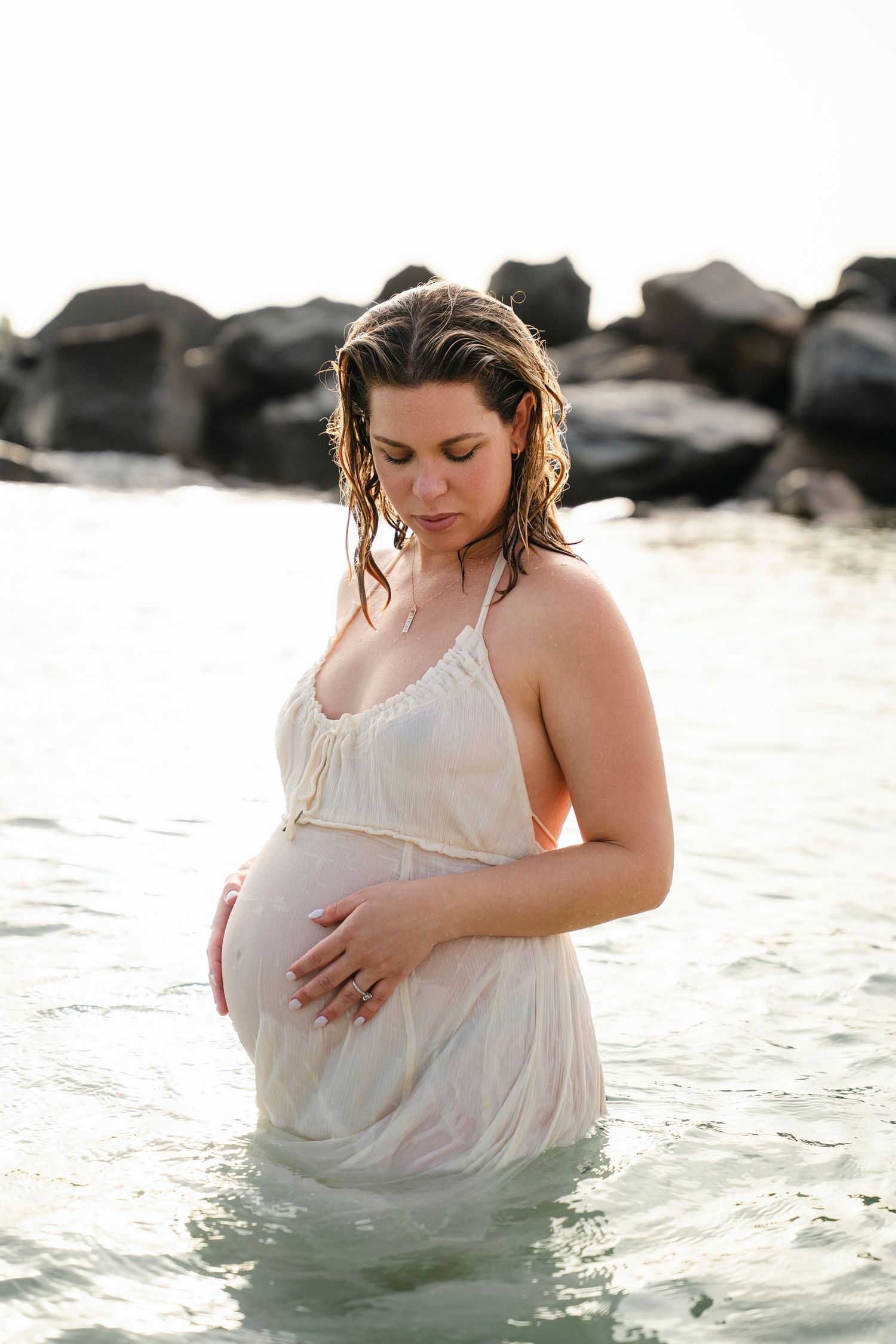 Maternity photoshoot in white dress at sunset by the ocean with rocks in background.