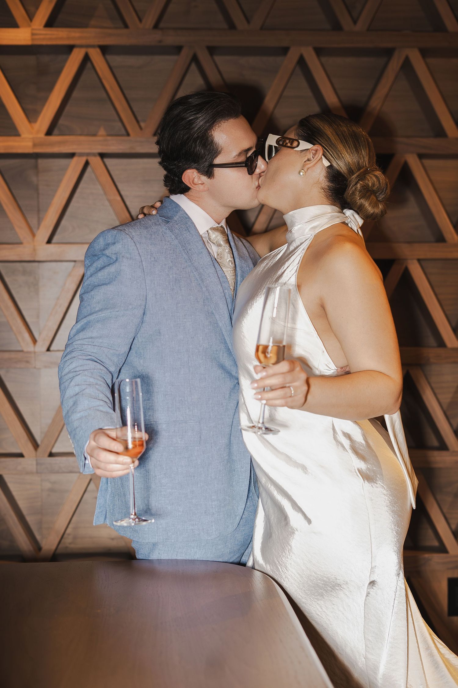 Couple in formal attire kissing while holding champagne glasses against a geometric wooden wall backdrop.