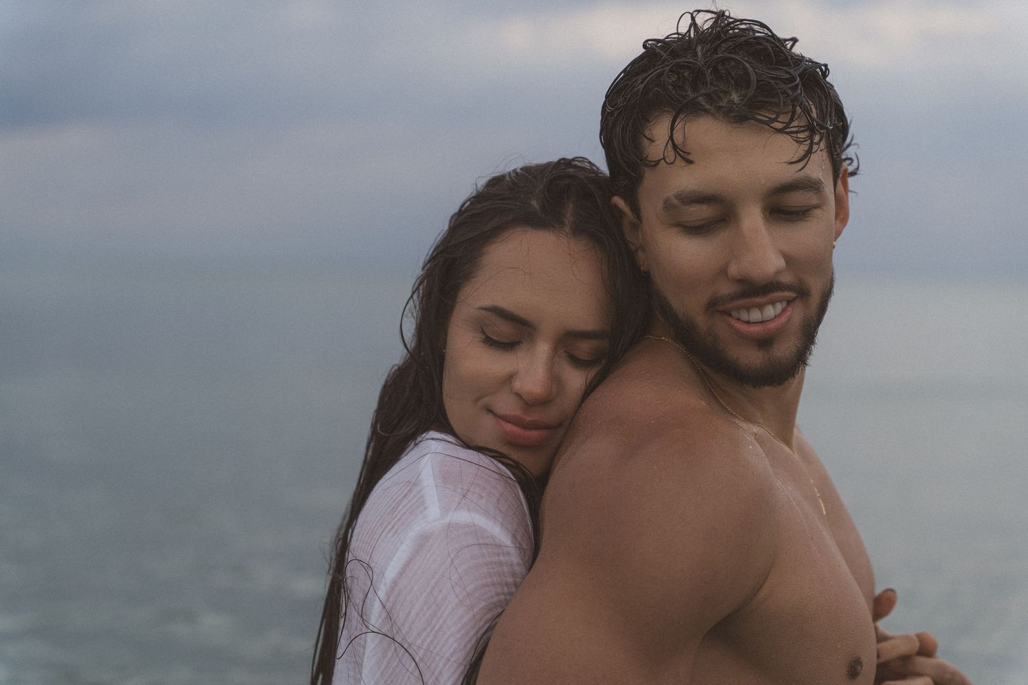 Romantic couple embracing on a beach, woman in white top resting head on man's shoulder near ocean.
