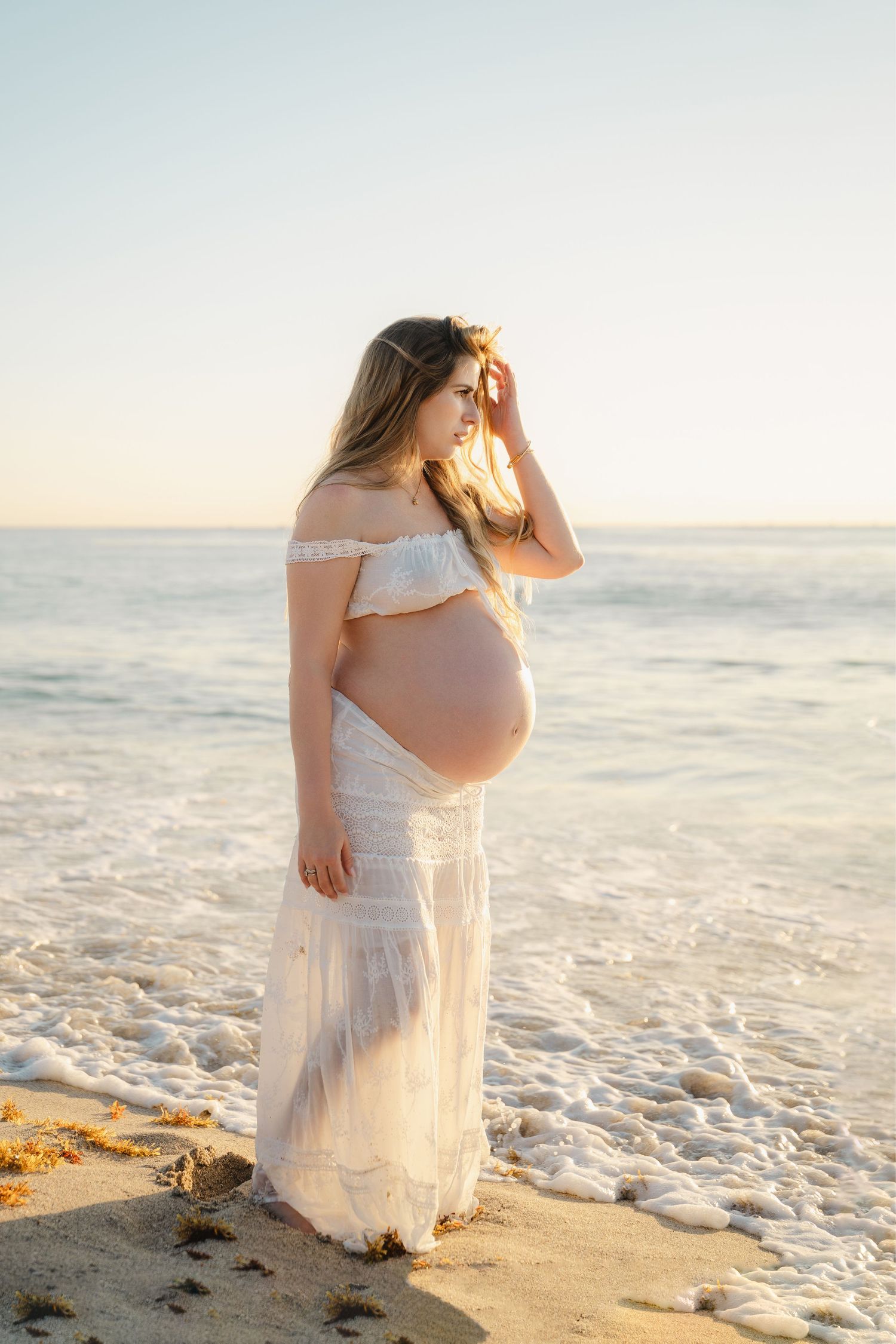 Pregnant woman in white off-shoulder dress standing on a sunny beach with ocean waves, golden hour maternity photo.