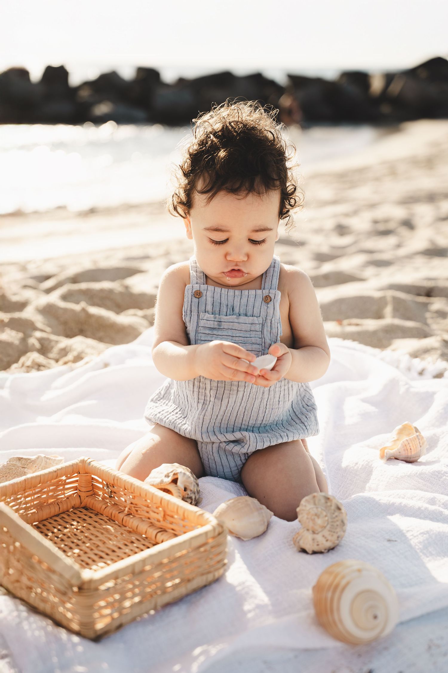 Small child in striped outfit plays with wooden basket on beach blanket.