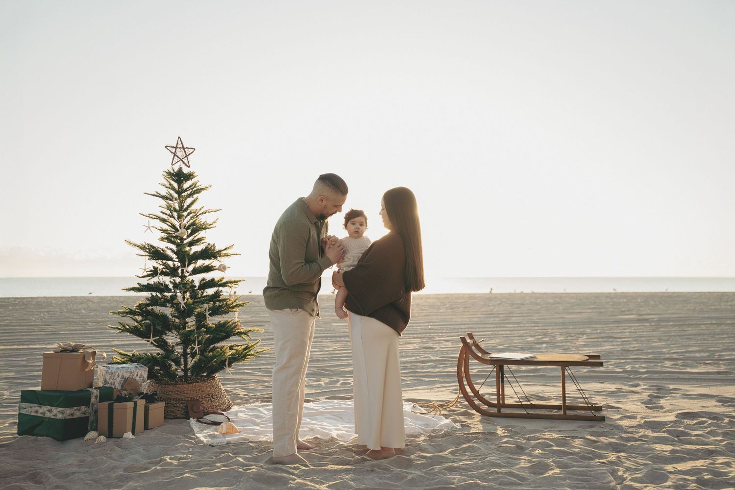 Family with baby celebrating Christmas on a sunny beach with a decorated tree and sled.