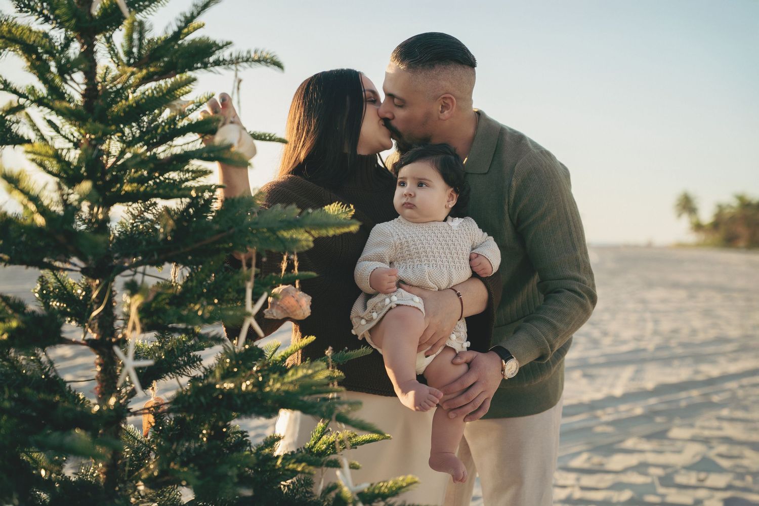 A couple kisses while holding their baby by a decorated Christmas tree on a sunny beach.
